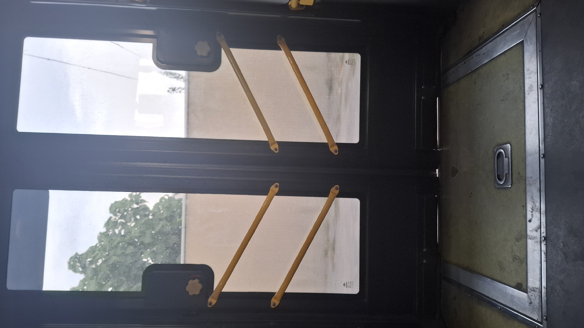 The image shows the interior of a dark-colored double door, likely from a bus or train, with large rectangular windows. There are yellow diagonal grab bars attached to the inside of each door panel. The floor in front of the doors is a worn, olive-green surface with metal trim around the edges and a recessed handle in the center. Outside the windows, a beige wall and greenery from trees are visible under a cloudy sky. The scene appears to be a public transportation vehicle, and the setting is not identifiable as a specific location within the city of Shymkent, Kazakhstan, based on the visual information provided. There are no people or discernible actions taking place in the image.