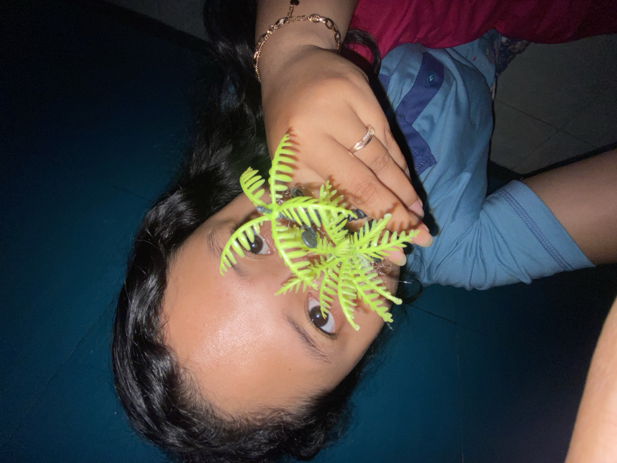 A person is holding a bright green, artificial plant up to their face, obscuring their mouth and nose. The person has dark hair and brown eyes. They are wearing a blue t-shirt with darker blue accents and a button closure. A ring is visible on their left hand, and a bracelet is on their right wrist. The background is a dark, teal-colored wall. The scene appears to be indoors, with some of a tiled floor visible in the lower left corner. The image does not contain information that allows for the identification of a specific location within the city of Gelam, Indonesia.