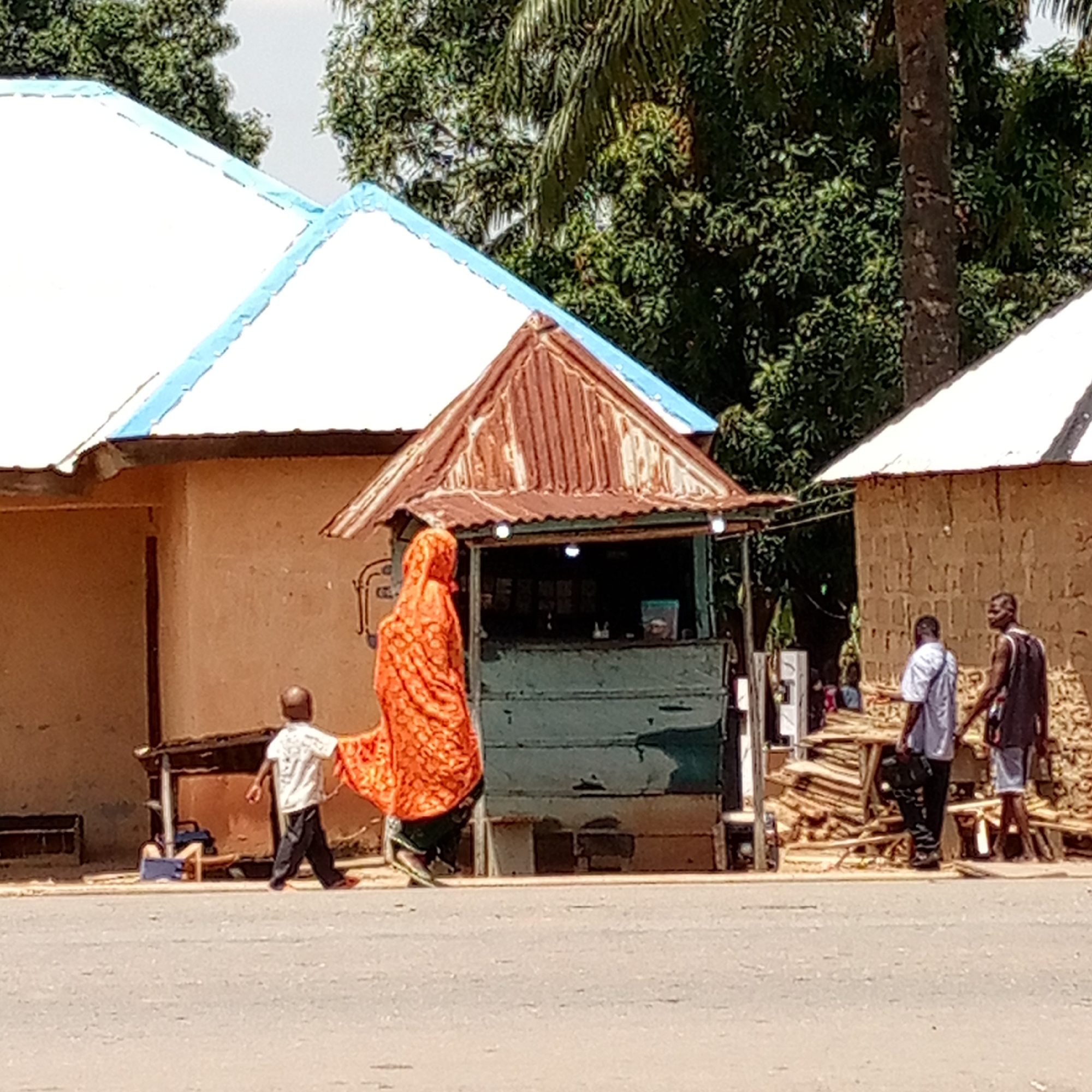 In Mararaba, Nigeria, an outdoor scene depicts two single-story buildings with white roofs edged in blue and natural brown walls. Between them is a small, green roadside stall topped with a rusty metal roof. An orange-robed woman and a young boy in a white shirt and dark pants stand next to the stall. Two men are standing by stacked wood piles next to the building to the right of the stall. The foreground is dominated by a paved road. The scene is set under bright daylight, with lush green trees visible in the background.
