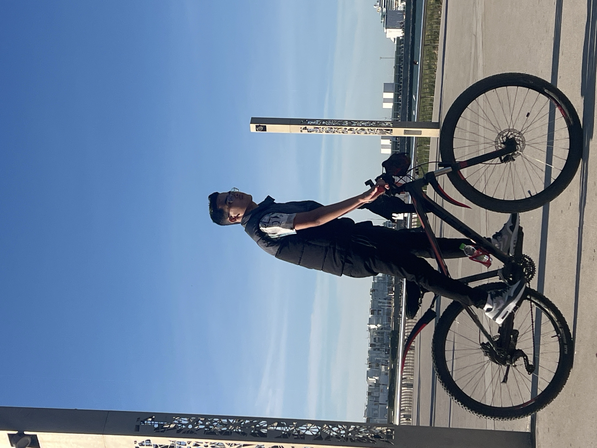 A young man stands with a bicycle in Tanger, Morocco. He is wearing glasses, a vest over a t-shirt, and pants with sneakers. The bicycle is a mountain bike with black and red accents. Behind him are buildings and a clear blue sky. There's an ornate metal structure and a pillar beside him.