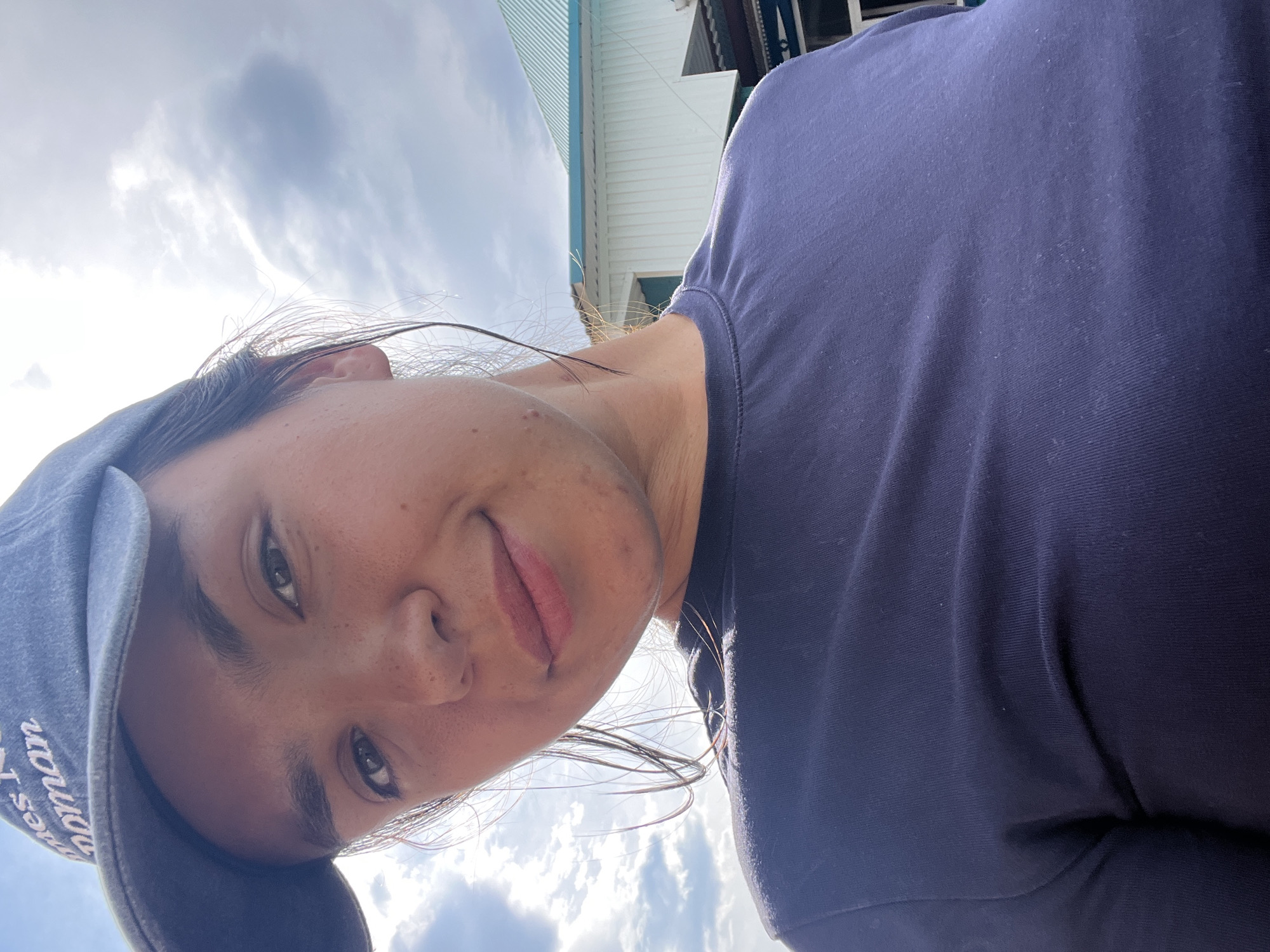 A young woman with brown hair pulled back, wearing a navy blue baseball cap and a navy blue t-shirt, is looking at the camera. Her cap has white lettering on it. She is outdoors with a cloudy sky and a building with a light-colored facade and a turquoise roof in the background. The image was taken at an angle looking up at her face, and part of her chest and shoulder are visible. She has freckles and a slight smile. It's not possible to determine the specific location within Oktyabrsky, Russia.
