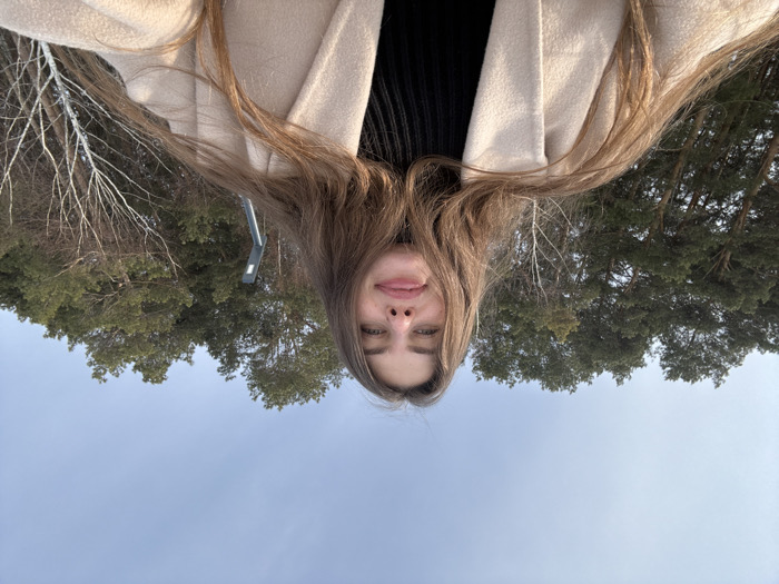 A low-angle frontal photograph captures a young adult female with long, wavy brown hair, centered in the frame. She wears a light beige overcoat over a dark, ribbed turtleneck. Her expression is characterized by a slightly open mouth with her tongue partially extended. The background consists of a clear, pale blue sky above a dense line of trees. Tall coniferous trees with dark green needles dominate the left and central portions of the background. To the right, several deciduous trees with light-colored, bare trunks and thin branches, consistent with birch trees, are visible. A dark, modern-style outdoor lamppost or light fixture is partially seen behind the woman's right shoulder, situated among the trees. The environment suggests an outdoor park or wooded area located within the city of Chaikovsky, Russia.
