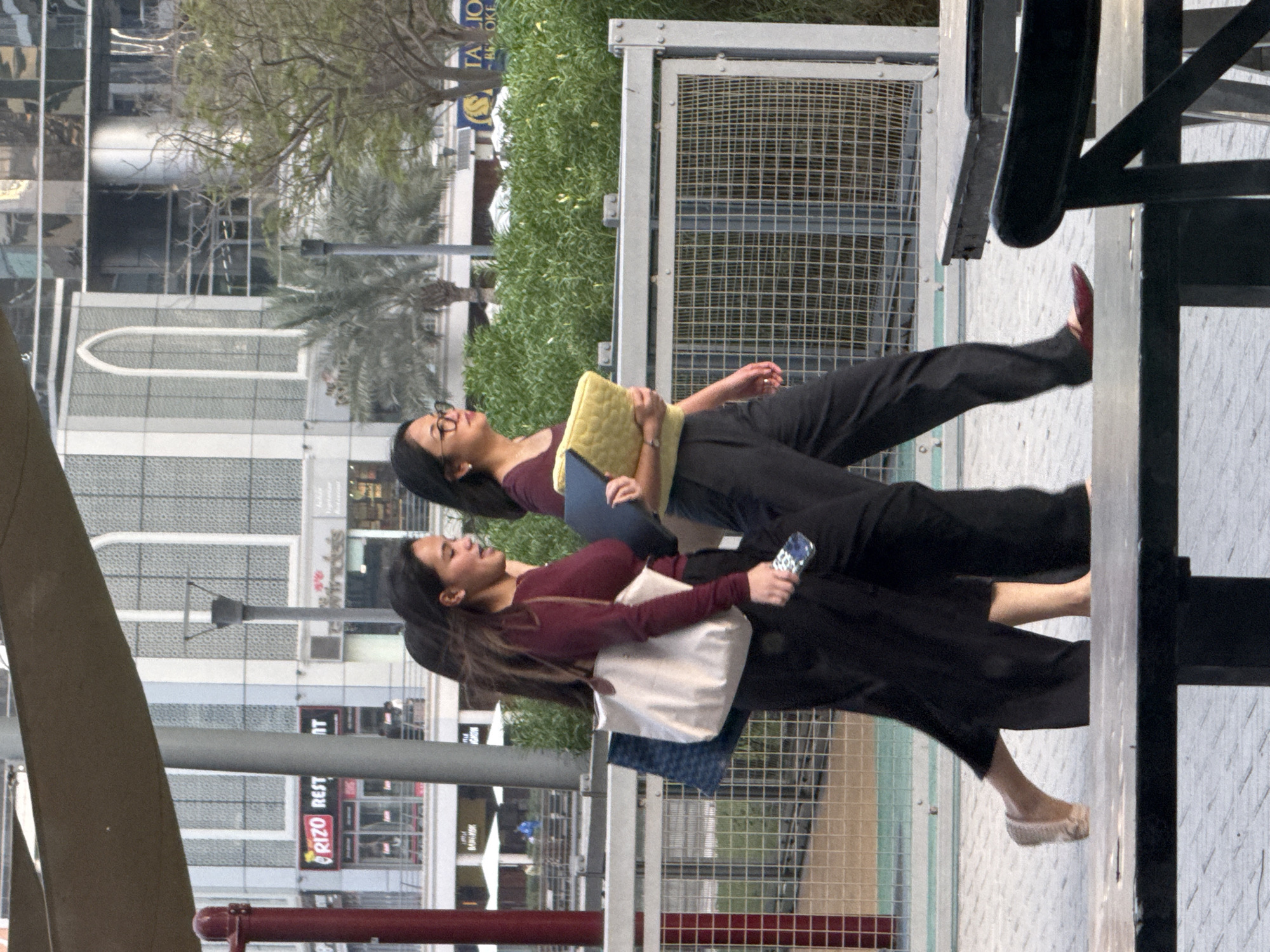 Two women are walking outdoors in the city of Dubai, United Arab Emirates, in front of a modern building with large windows and balconies, and green trees. The woman on the left is wearing a dark maroon long-sleeved shirt and black pants, and carrying a large white tote bag with a blue and white patterned bag inside. She has long dark hair and is wearing light-colored flats. The woman on the right is wearing a dark maroon long-sleeved shirt and black pants, and is holding a yellow padded item and a dark blue folder. She has long dark hair and is wearing glasses and black flats. They are walking on a paved walkway next to a metal fence. In the background, there are several shops and restaurants, including one with a sign that says "RIZO REST".
