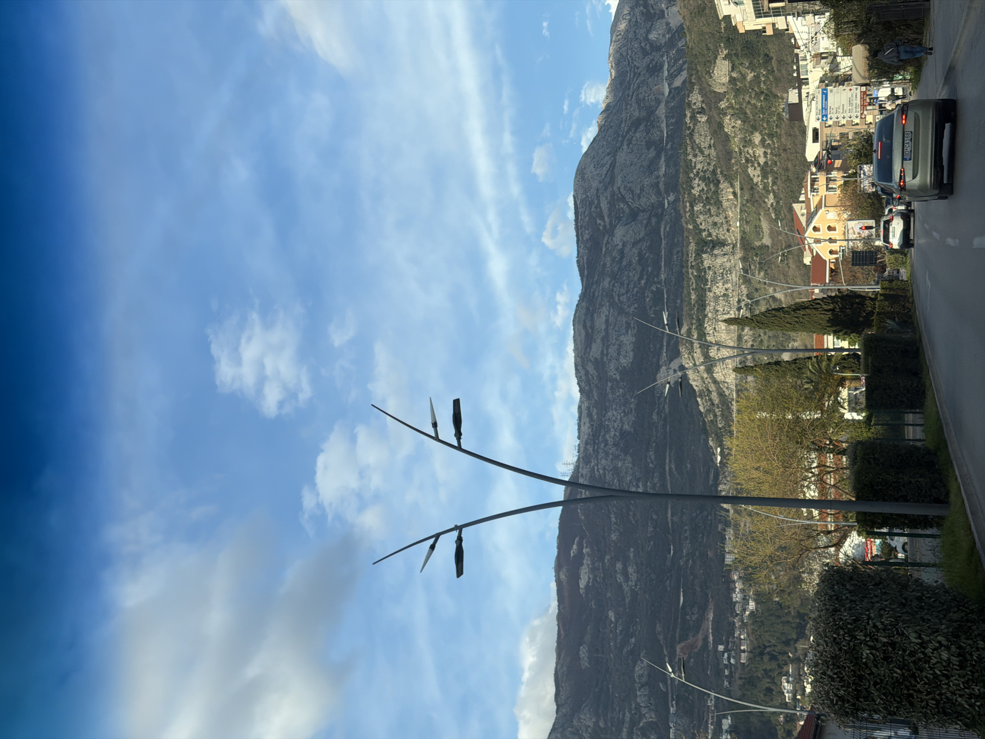 The image shows a view in Budva, Montenegro, featuring a large mountain range on the right side with visible geological features. The sky is blue with streaks of white clouds on the left. A modern street light with two metal branches extends into the frame. A road with cars, including a silver SUV with Montenegrin license plates, runs along the right side of the image. Buildings and trees are visible between the road and the mountain.