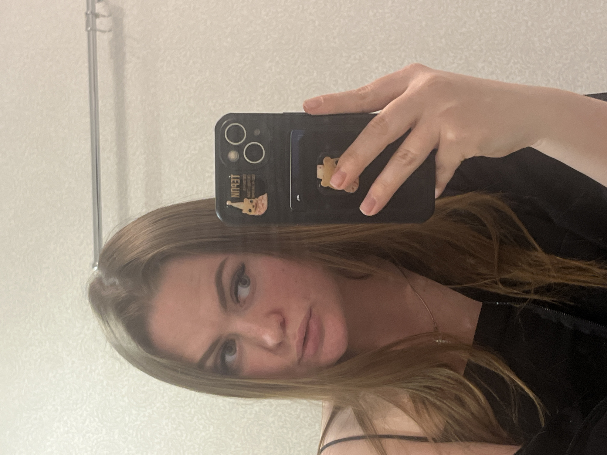 This image is a mirror selfie, rotated 90 degrees clockwise. It features a person, likely female, with light brown or blonde hair, fair skin, and full lips, looking towards the left of the frame. Her eyes are light-colored, and she has visible eyelashes and shaped eyebrows. She wears a dark top, with a thin strap visible over her right shoulder, and a delicate silver necklace around her neck. Her expression is neutral, with a slight pout.
The person is holding a black smartphone in her right hand, which is reflected in the mirror. The phone appears to be an iPhone, identifiable by its dual camera lenses on the back. It is encased in a clear or translucent case. Attached to the back of the case is a small, light-colored pop socket or phone grip, which the person's fingers are holding. A small card holder is also visible on the back of the case. On the lower left of the phone case, below the camera lenses, there is a sticker or design featuring a cartoon character, possibly a small animal in a hat. The mirrored text "TEBUN" is clearly visible on the phone case, positioned horizontally below the camera lenses.
The background visible in the mirror reflection is a light-colored wall with a subtle, textured pattern, resembling wallpaper. A thin, vertical metal rod or rail is visible on the left side of the reflection, extending from the top edge. The setting appears to be indoors, possibly a room or bathroom, under soft, even lighting.