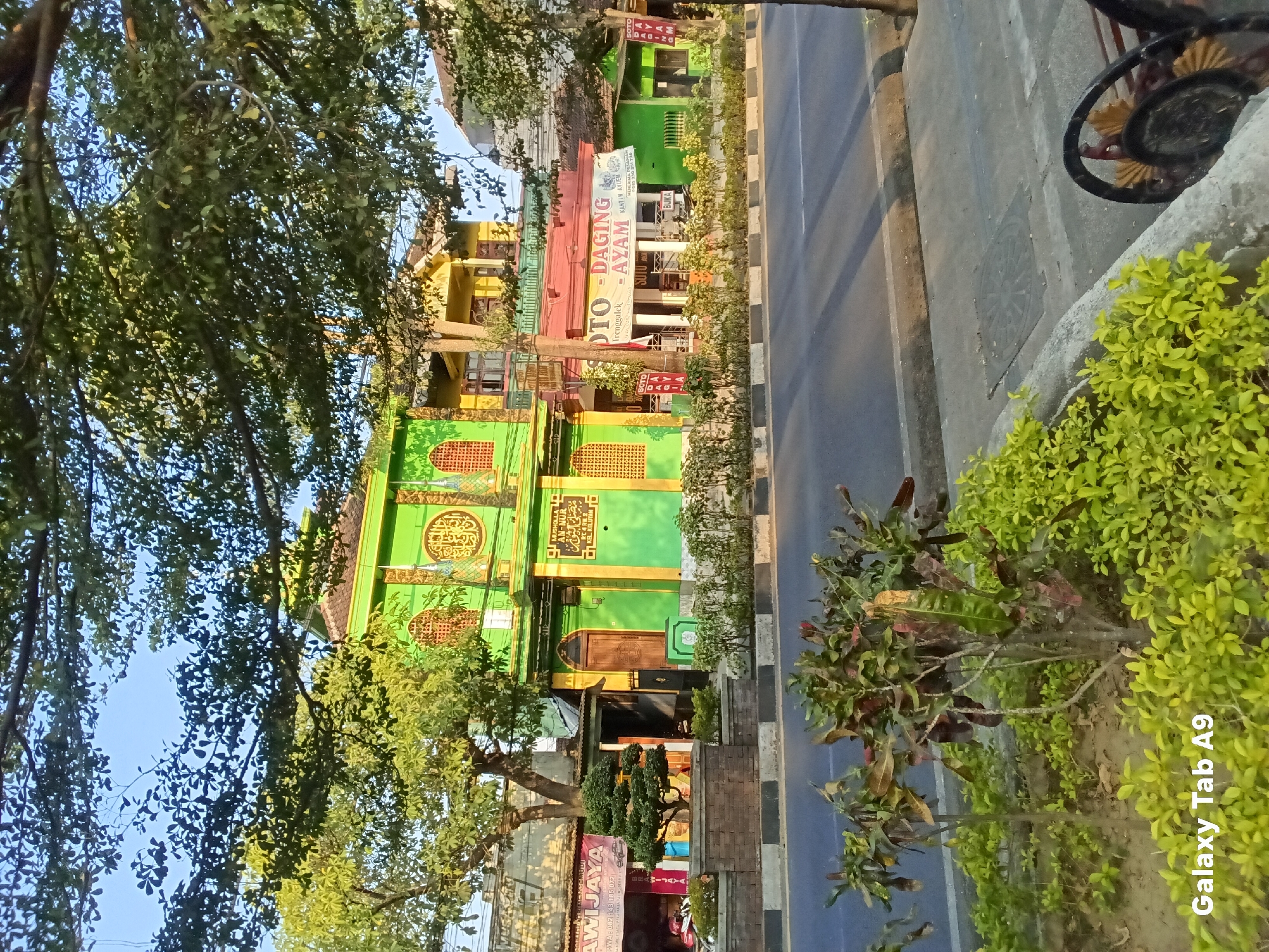 The image shows a street scene in Kelutan, Indonesia. On the left is a bright green and yellow building with intricate window designs and Arabic calligraphy. A sign on this building reads "MUSHOLLA AL-MU'MIN" and "KEL. KELUTAN". To the right of this building, there is a yellow building and a red awning above a restaurant. A banner on the awning reads "SOTO - DAGING - AYAM", indicating it serves meat and chicken soup. Further to the right is a smaller green building. In front of the buildings, there is a sidewalk with a low brick wall and decorative plants, including a bonsai tree. A paved road with a double white line runs in front of the sidewalk. Trees with green foliage overhang the scene, and the sky is clear and blue. The lighting suggests it is daytime, likely morning or late afternoon due to the warm tones and shadows.