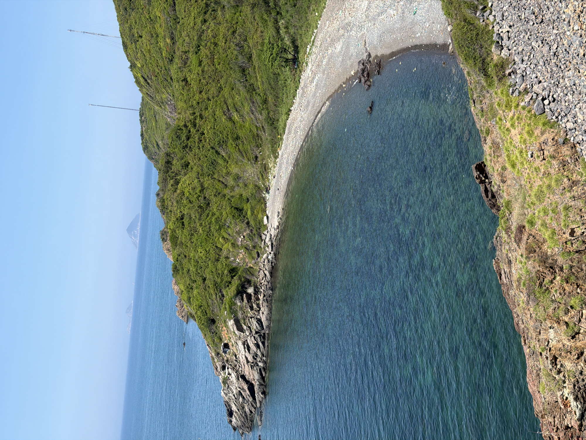This high-angle photograph depicts a coastal landscape in Bac Nha Trang Ward, Vietnam, under a clear, bright blue sky. The expansive sea is calm, displaying a transition from deeper blue in the distance to a translucent turquoise closer to the shore, where submerged features are visible. Three small islands are discernible on the distant horizon.
The coastline on the left features a rugged, rocky promontory of brown and grey stratified rock, extending into the water. This formation helps define a crescent-shaped pebble beach situated at the base of a steep, densely vegetated hill. The beach consists primarily of light-colored pebbles, with a visible accumulation of debris along the line where it meets the land. A solitary small, green-leafed tree stands towards the rear of the beach, positioned slightly to the right. The hillsides are covered in lush, dense green foliage. On the summit of the hill to the right, two tall, slender communication or observation towers are prominently visible against the sky. The immediate foreground on the lower right shows a rocky slope interspersed with sparse dry grass and a field of loose grey gravel. No individuals or active human interventions are depicted in the scene.