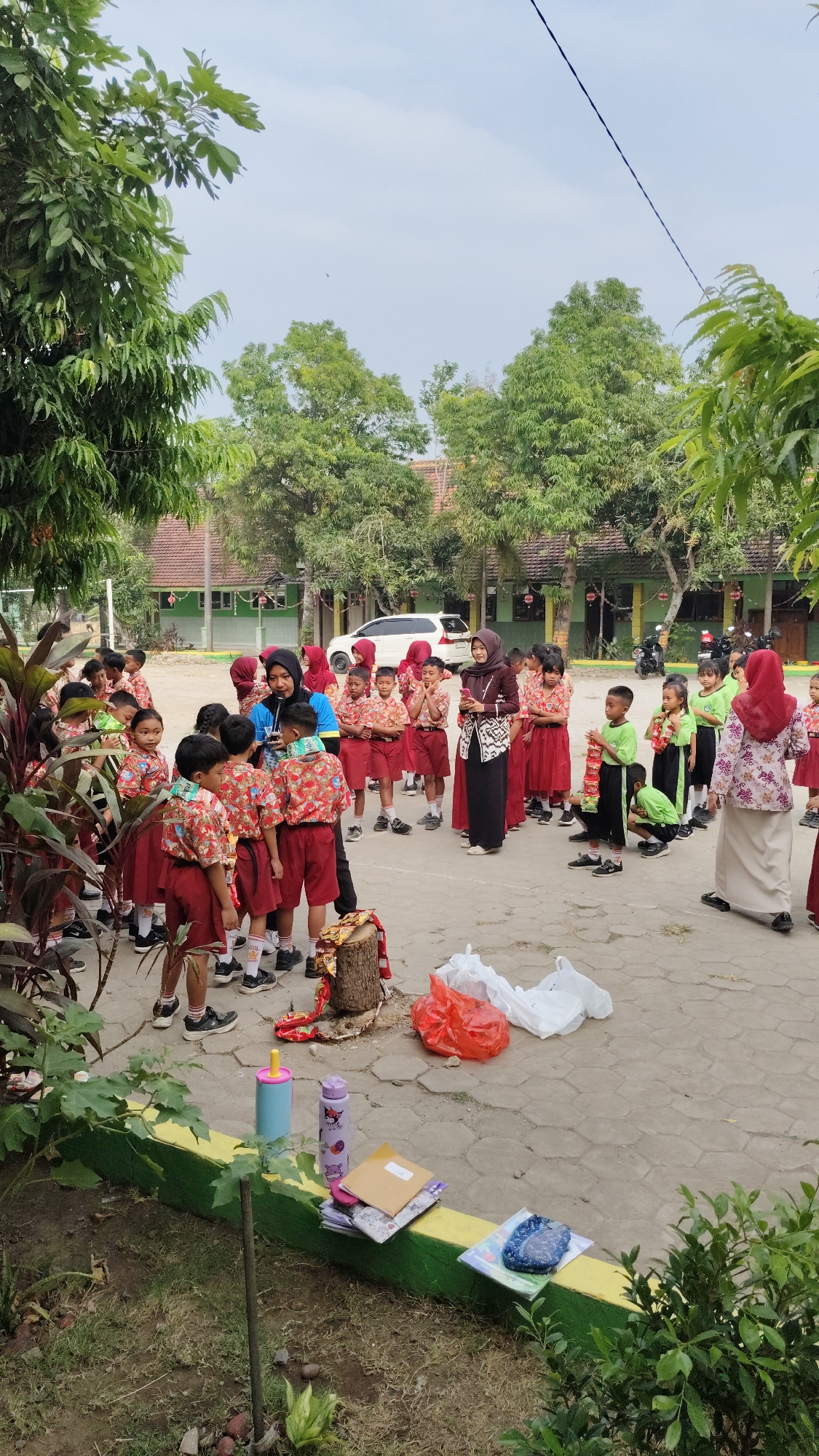 In Banjarsari, Indonesia, a group of children in school uniforms are gathered outdoors, possibly for an activity or lesson. The children, mostly in red and white uniforms, are listening to two adult women who appear to be teachers. One teacher, wearing a hijab and a brown patterned outfit, is holding a phone, while another, in a blue shirt, is speaking into a microphone. The scene is set in a schoolyard with paved ground and trees. Several objects, including plastic bags, a log with decorative items, and books, are scattered around the area, indicating a potential event or outdoor classroom setup.