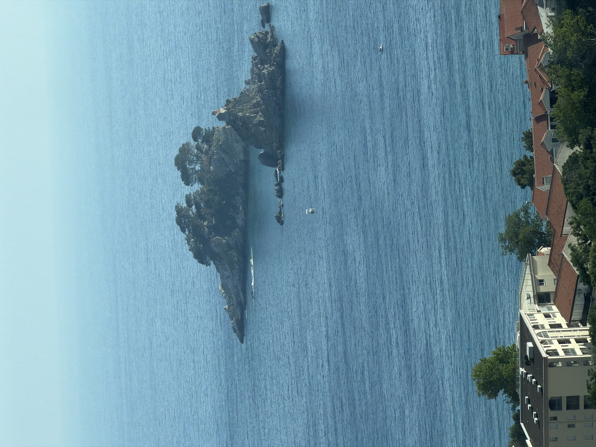 The image displays a vibrant coastal landscape under bright daylight, likely depicting Stupovi, Montenegro, with a prominent rocky islet. The central focus is a rugged, tree-covered island emerging from the clear blue sea. On the island, a small, red-roofed building, possibly a chapel or house, is visible among the greenery. Smaller rock formations extend from the main island into the water.
The expansive sea fills most of the frame, showing visible ripples and textures on its surface, indicating gentle movement. Two small white boats are present on the water, one closer to the islet and another further out in the sea. Along the right side of the image, the coastline features several buildings with distinctive red-tiled roofs, characteristic of Mediterranean architecture. These structures are surrounded by green trees, suggesting a residential or resort area. The sky above is a clear, light blue, without clouds, emphasizing the sunny weather.
No specific human activity is discernible, and no visible text appears in the image. The scene conveys a sense of tranquility and natural beauty, typical of a coastal destination.