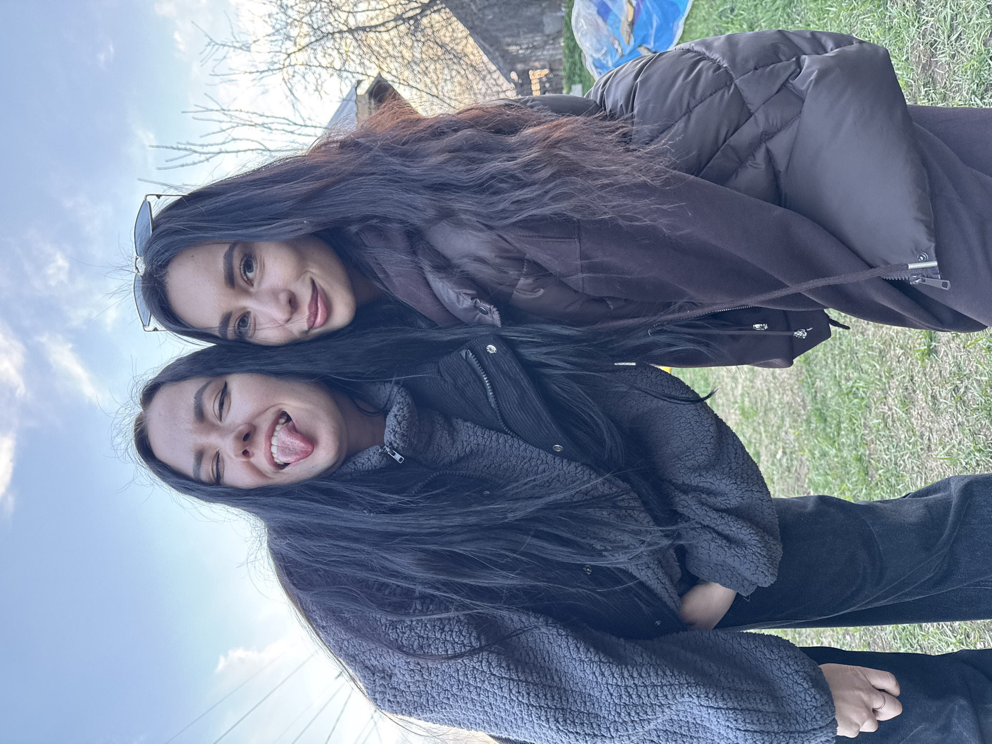 Two women are posing outside in an unidentified outdoor location, potentially in the city of Chaikovsky, Russia. The woman on top is wearing sunglasses and a brown puffer jacket. The woman beneath her is sticking her tongue out. She is wearing a gray zip-up jacket and black pants. The environment includes grass, trees, a building in the background, and a blue sky with clouds.