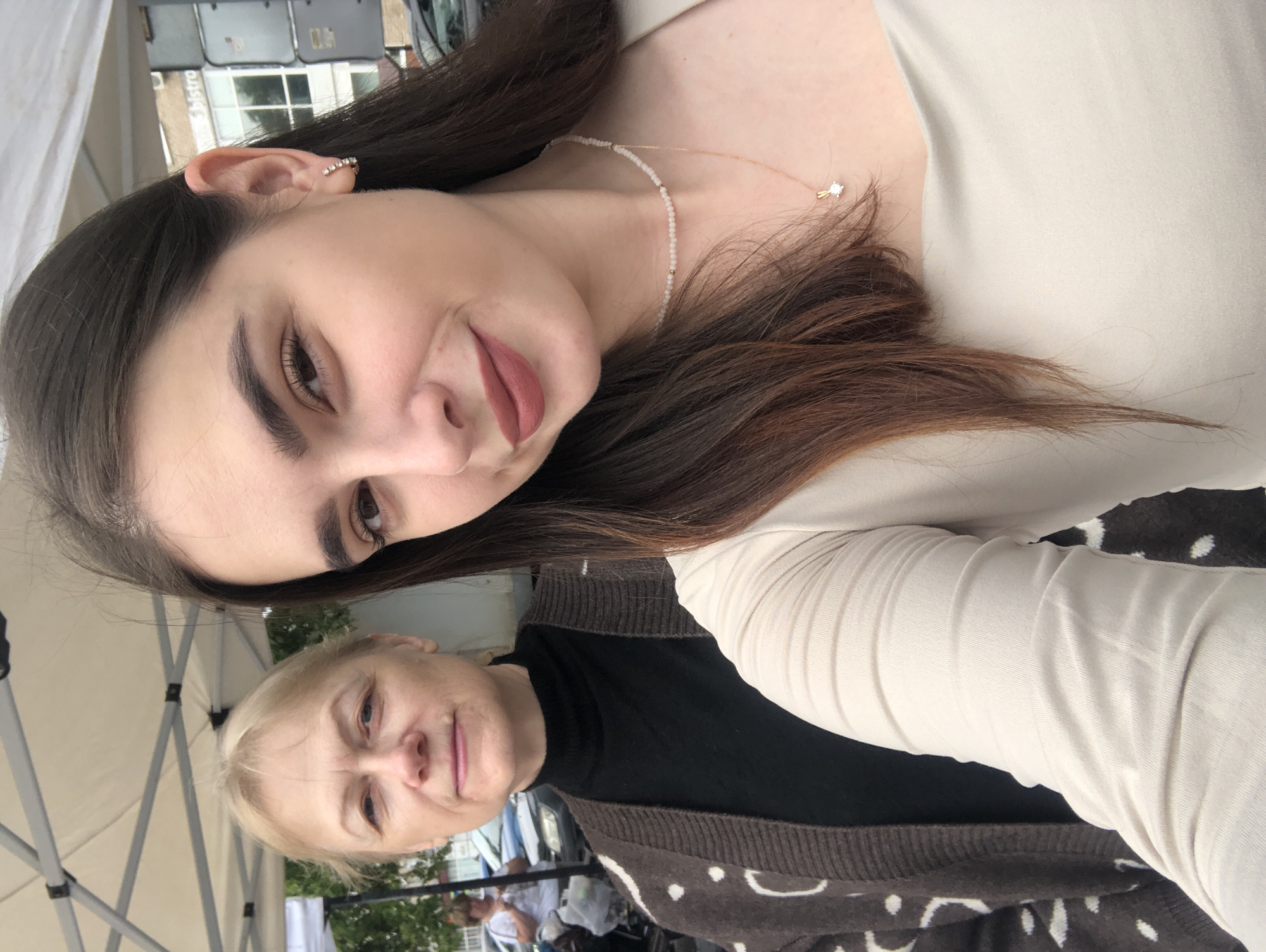 The image shows a selfie taken by a young woman, with an older woman visible behind her. They are standing outdoors, under a white canopy that appears to be part of a market stall or event tent. The background shows a street scene with cars and a building with a sign that reads "Bistro". The young woman is wearing a light-colored top, a pearl necklace, and a pendant. The older woman is wearing a black turtleneck and a brown cardigan with a white dotted pattern. Both individuals appear to be casually dressed. The lighting suggests it is daytime. The specific location in Chaikovsky, Russia, cannot be determined from the visual information in the image.