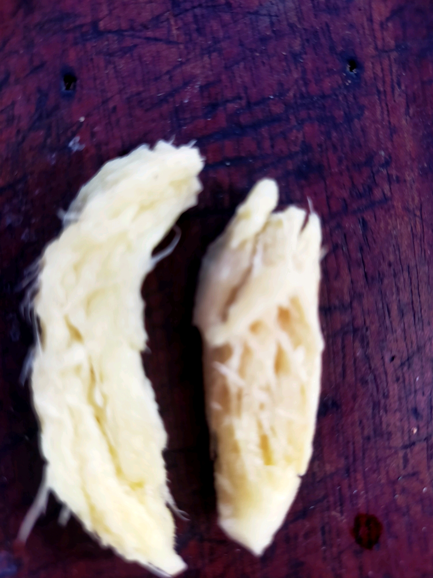Two pieces of shredded yellow material are laid out on a dark, textured surface that appears to be wood. The material on the left is long and stringy, resembling shredded coconut or finely pulled meat. The piece on the right is shorter and appears to have a more fibrous, textured interior, with some lighter-colored strands visible. The surface beneath them is a deep reddish-purple, with visible grain and some darker specks or marks. The lighting is soft and diffused, with no harsh shadows. The image is slightly out of focus, particularly at the edges. This image does not contain any identifiable information related to a specific location within the city of Surabaya, Indonesia.