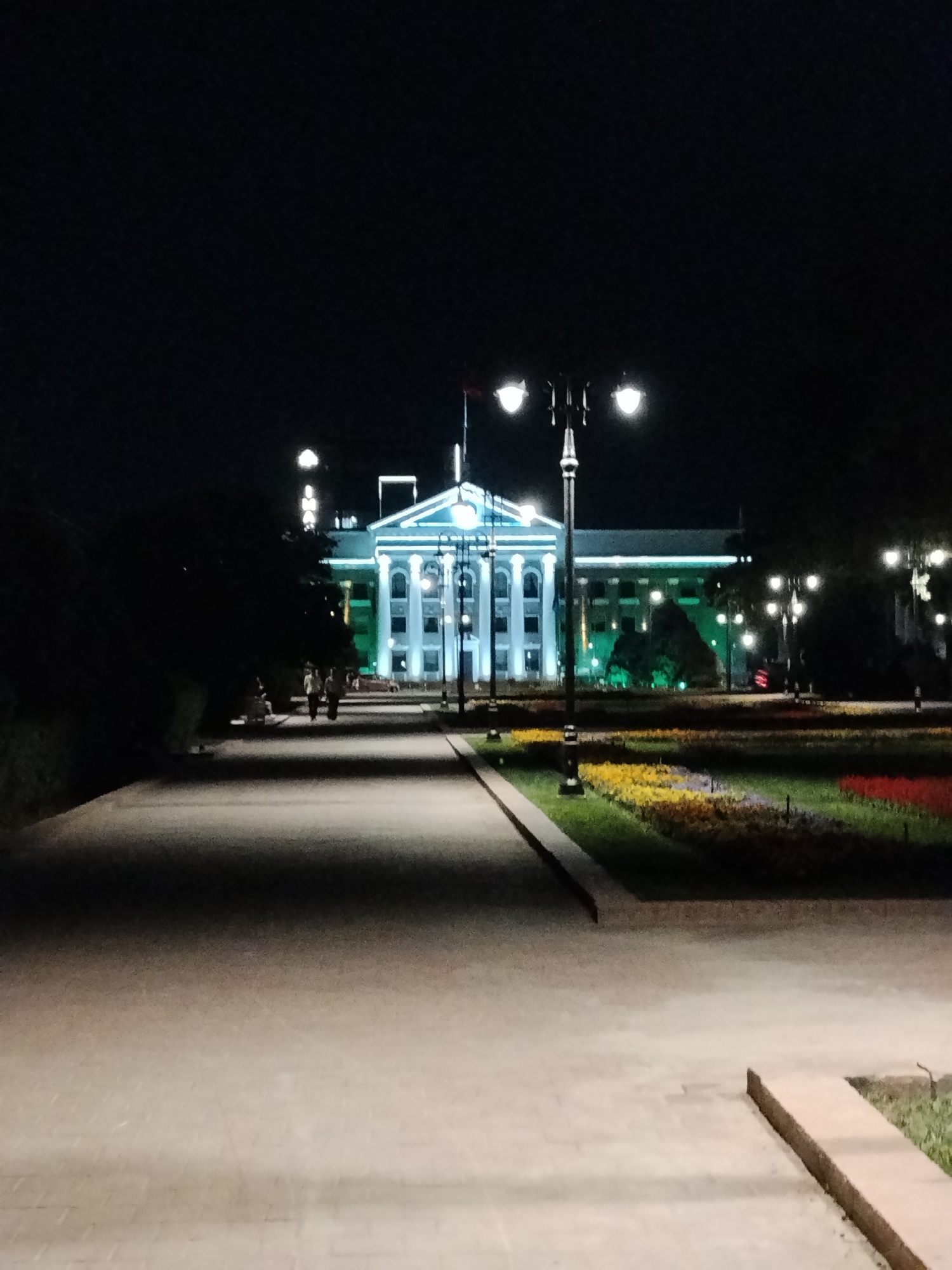 A wide-angle, long-exposure nighttime photograph captures the illuminated facade of the State Historical Museum in Bishkek, Kyrgyzstan. The building is a grand, neoclassical structure, lit with cool blue and green lights that highlight its architectural features. A large, ornate lamppost stands in the foreground on the left, its two globes casting warm light onto a paved walkway. To the right of the lamppost, a flower bed is visible, planted with rows of yellow, red, and green flowers. Several people are walking along the pathway towards the museum, their figures blurred by the long exposure. The sky is dark and starless, and the surrounding trees are silhouetted against the building's light. The overall impression is one of quiet grandeur and a peaceful urban evening.