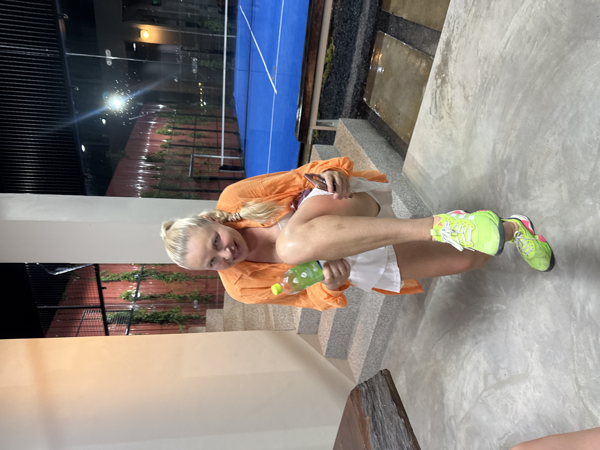 A woman with blonde hair in a ponytail, wearing an orange open shirt over a white top and skirt, and neon green athletic shoes, kneels on a concrete floor. She is holding a green bottle in her right hand and a mobile phone in her left hand. She is smiling at the camera. In the background, a blue sports court with white lines is visible, suggesting an outdoor or semi-outdoor facility, possibly a padel or tennis court. The area appears to be a modern, possibly resort-style, location, given the architectural style of the stairs and the fencing behind the court. The lighting suggests it is evening or night. Based on the provided context, the location is in the city of Ko Samui District, Thailand.