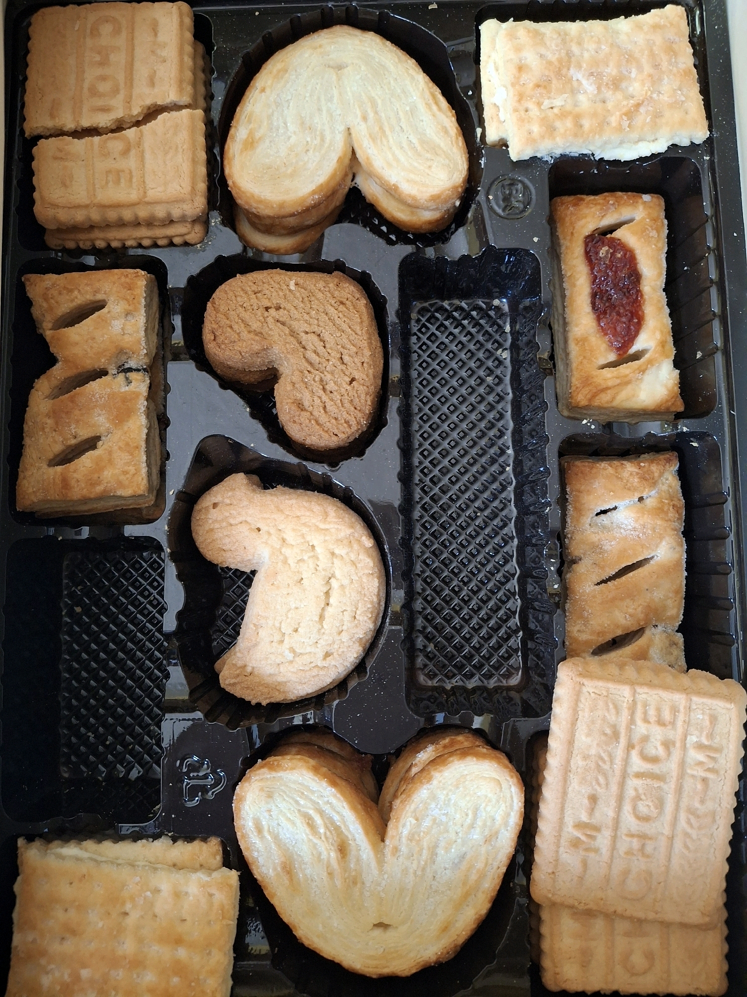 The image shows a black plastic tray filled with an assortment of baked goods, resembling a cookie or pastry assortment. The tray is divided into compartments. In the top left, there are stacked rectangular cookies with embossed lettering that appears to read "M CHOICE". To their right are several layered, butterfly-shaped pastries, also known as palmiers. Below the rectangular cookies, in the left column, are two more rectangular pastries with visible slits on top, one of which has a filling visible, possibly fruit jam. In the center of the tray, there is a heart-shaped cookie, and below it, another pastry that resembles a flattened, irregular-shaped cookie. In the right column, starting from the top, there is a rectangular pastry with a white filling, followed by another rectangular pastry with slits and a jam filling. The bottom right compartment contains stacked rectangular cookies that appear identical to those in the top left, also embossed with "M CHOICE". The tray itself is made of black plastic with a textured surface in some compartments. The baked goods are golden brown and appear to be of various textures, some flaky and layered, others more solid and crumbly. There are no people or animals in the image, and the environment appears to be a studio setting or a clean, neutral background, without any discernible geographical markers or elements that would suggest a specific location like Lumajang, Indonesia.