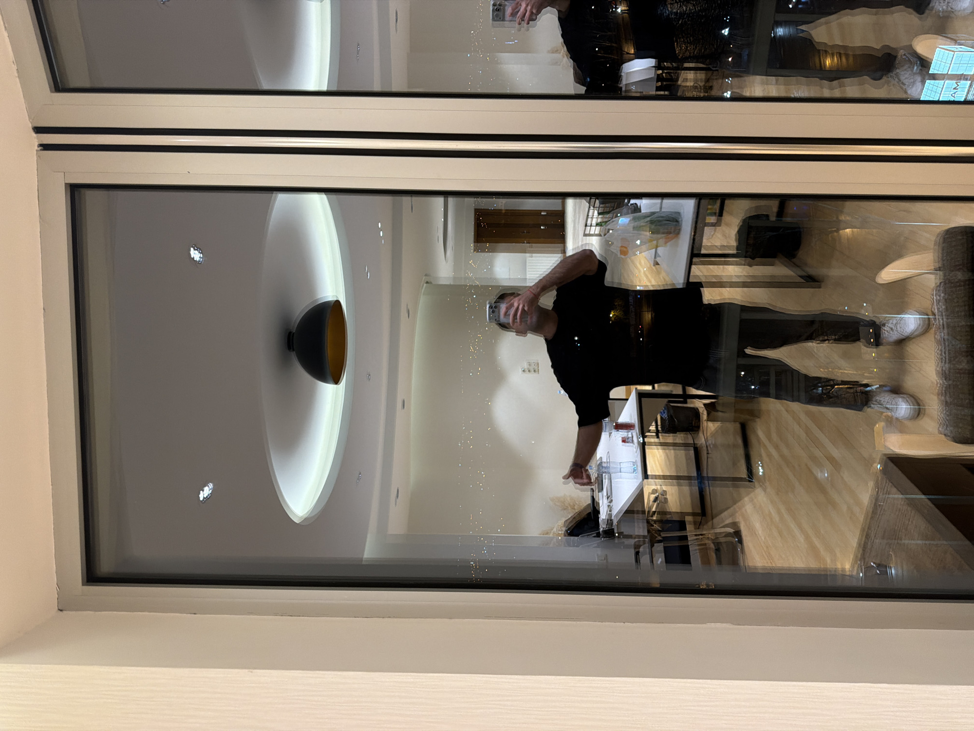 A person is reflected in a window, holding a phone and taking a picture. The reflection shows the interior of a room with a modern ceiling light and the exterior showing city lights at night. The location is Tbilisi, Georgia. Furniture and decor are visible in the reflection.