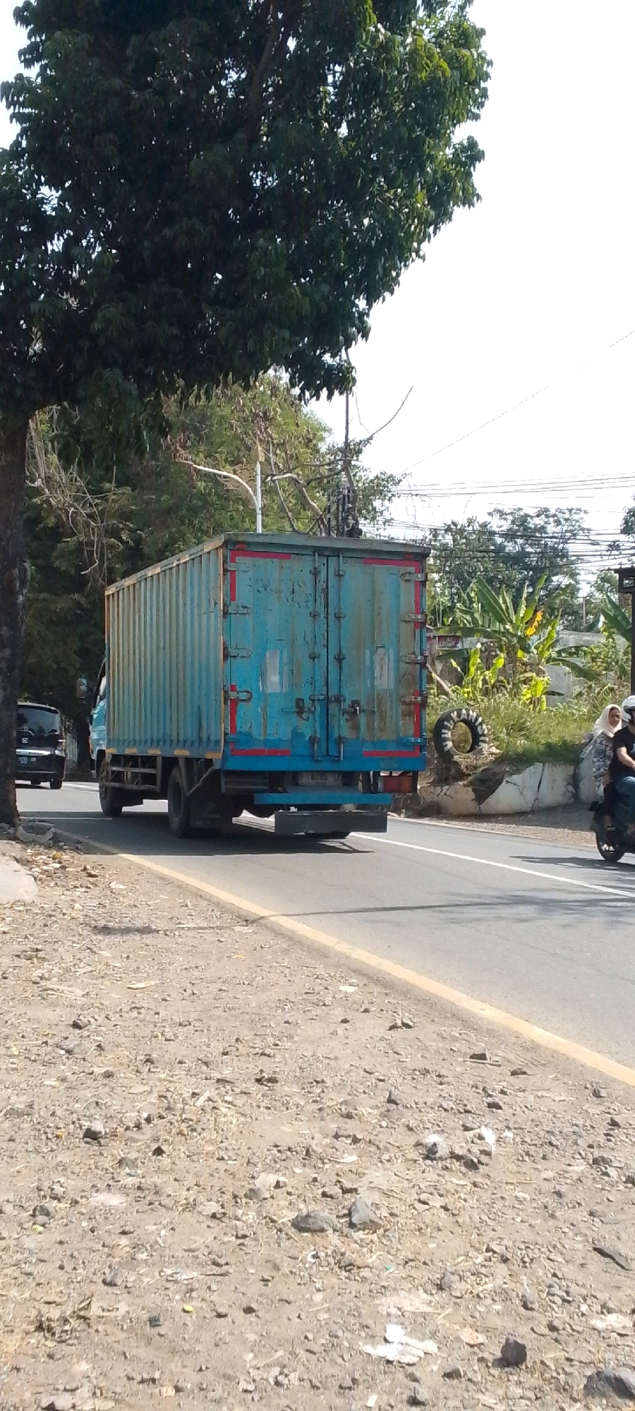 A blue cargo truck with a weathered, somewhat rusty exterior is traveling on a paved road in Cilaku, Indonesia. The truck's cargo container is ribbed vertically and has two rear doors with horizontal red and blue trim lines. The truck has dual rear wheels on the right side, and the front is partially visible. To the left of the truck, a black SUV is also moving down the road, partially obscured by a large tree with dark green foliage that dominates the left side of the frame. The road has a yellow line marking the edge on the right. On the right side of the road, there is a motorcycle with two occupants, a person driving and a passenger wearing a light-colored headscarf. Behind them, there is a pile of debris including a deflated tire and some broken concrete. The environment appears to be a semi-rural or suburban area with trees and vegetation visible in the background, under a bright, clear sky. The ground along the side of the road is unpaved and covered with gravel, dirt, and scattered debris.