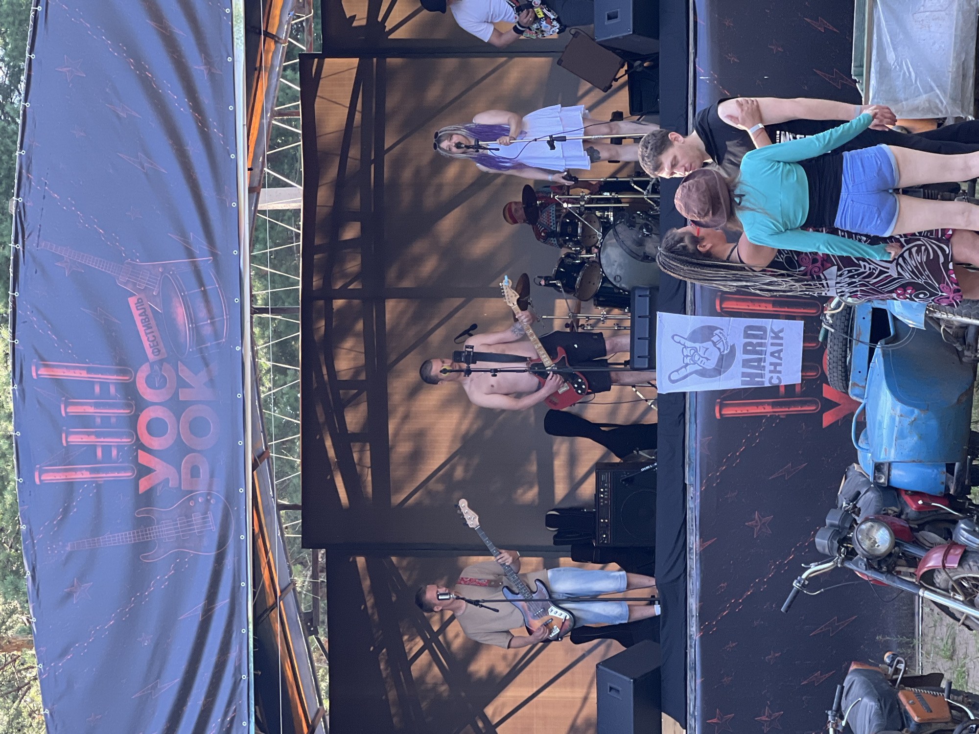 A rock band performs on an outdoor stage. The band members include a male guitarist in a tan shirt and denim shorts, a shirtless male guitarist, a female singer with purple hair in a white dress, and a male drummer. In the foreground, three people are interacting: a woman with braided hair, a woman wearing a turquoise cropped shirt and denim shorts, and a man in a black t-shirt. Two motorcycles and a blue sidecar are parked in front of the stage. A banner behind the band reads "YOC POX FESTIVAL". The scene appears to be taking place in the city of Chaykovsky, Russia, likely during a summer event.