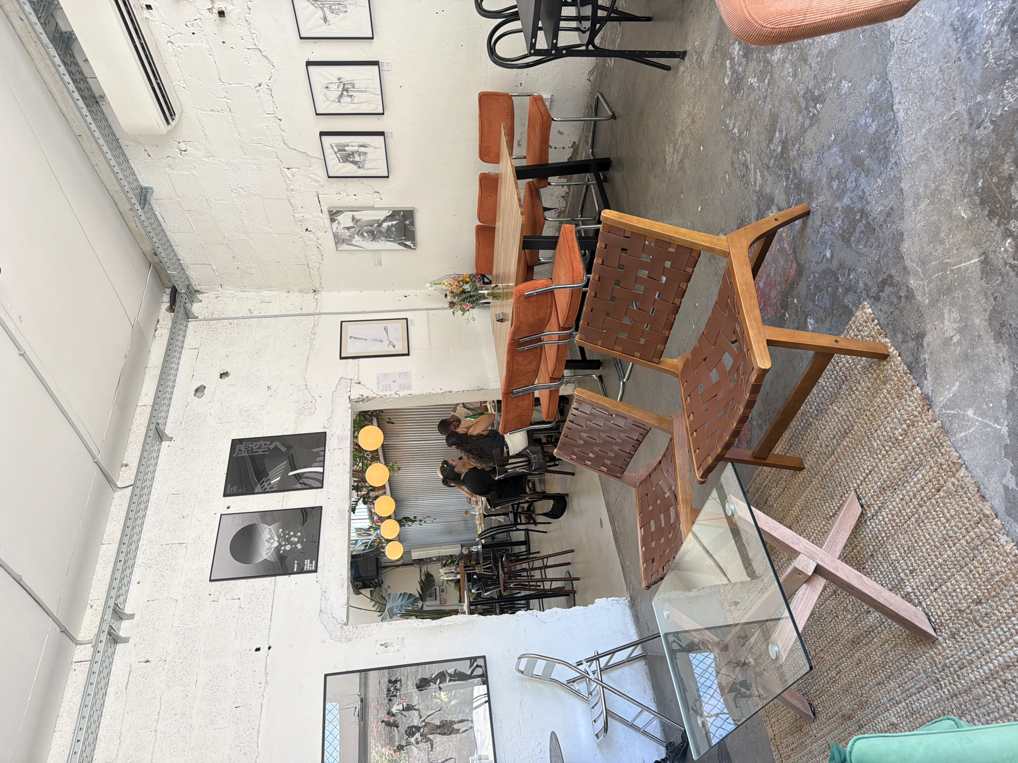 The image depicts the interior of a cafe in Tel Aviv, Israel. The space features exposed brick walls, concrete floors, and industrial-style lighting. In the foreground, a glass-topped coffee table with a wooden base sits on a woven rug, accompanied by two stylish wooden chairs with woven brown leather seats and backs.
Further into the cafe, a long wooden dining table is surrounded by orange upholstered chairs. A vase of flowers is placed on the table. The back wall displays a series of framed black and white artworks, including sketches and graphic prints. An air conditioning unit is mounted high on the wall.
Through an open doorway, a seating area with patrons is visible, along with more tables and chairs. Yellow spherical lights hang from the ceiling in this area, and tropical plants add a touch of greenery. The overall ambiance suggests a trendy and relaxed atmosphere.