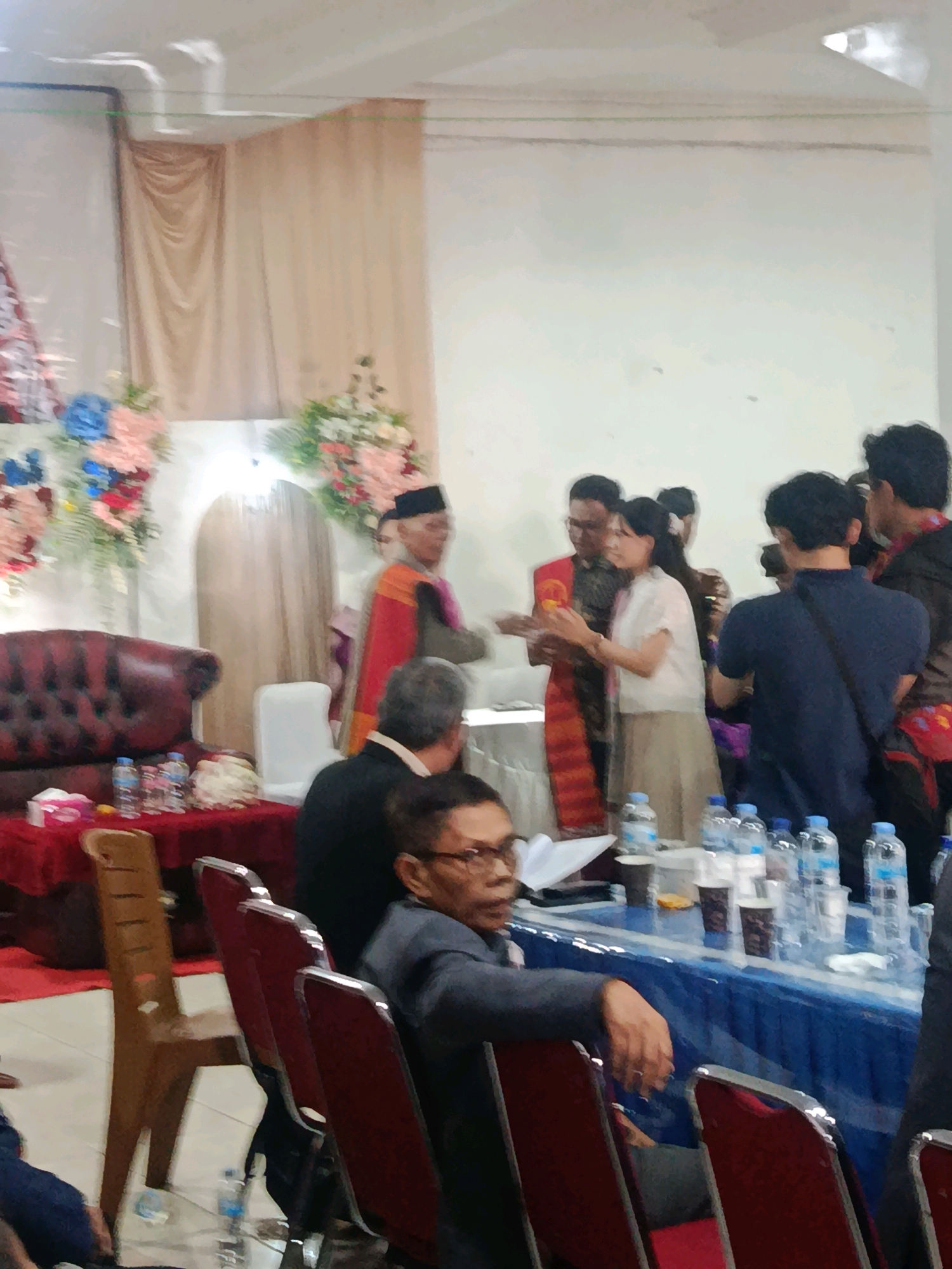 The image depicts an indoor event, likely a ceremony or formal gathering, possibly in Babelan, Indonesia. In the background, light brown curtains hang from the ceiling, flanking a doorway with a light-colored, arched door. To the left, a floral arrangement features pink, blue, and white flowers, and to the right, another arrangement includes pink, white, and green flowers. A prominent dark red leather sofa is situated in the left mid-ground.
In the center of the image, several individuals are interacting. A man in traditional attire, a red and orange sash over a dark outfit and a black cap, stands near the doorway. To his right, a woman in a white blouse and a beige skirt is engaged in conversation with him and another man. Other attendees are visible in the foreground and mid-ground, seated in chairs or standing. A table draped with a blue cloth is in the foreground, laden with bottles of water, cups, and tissues. A man in the right foreground is seated, facing left, wearing glasses and a dark suit. The overall lighting suggests an indoor event space.