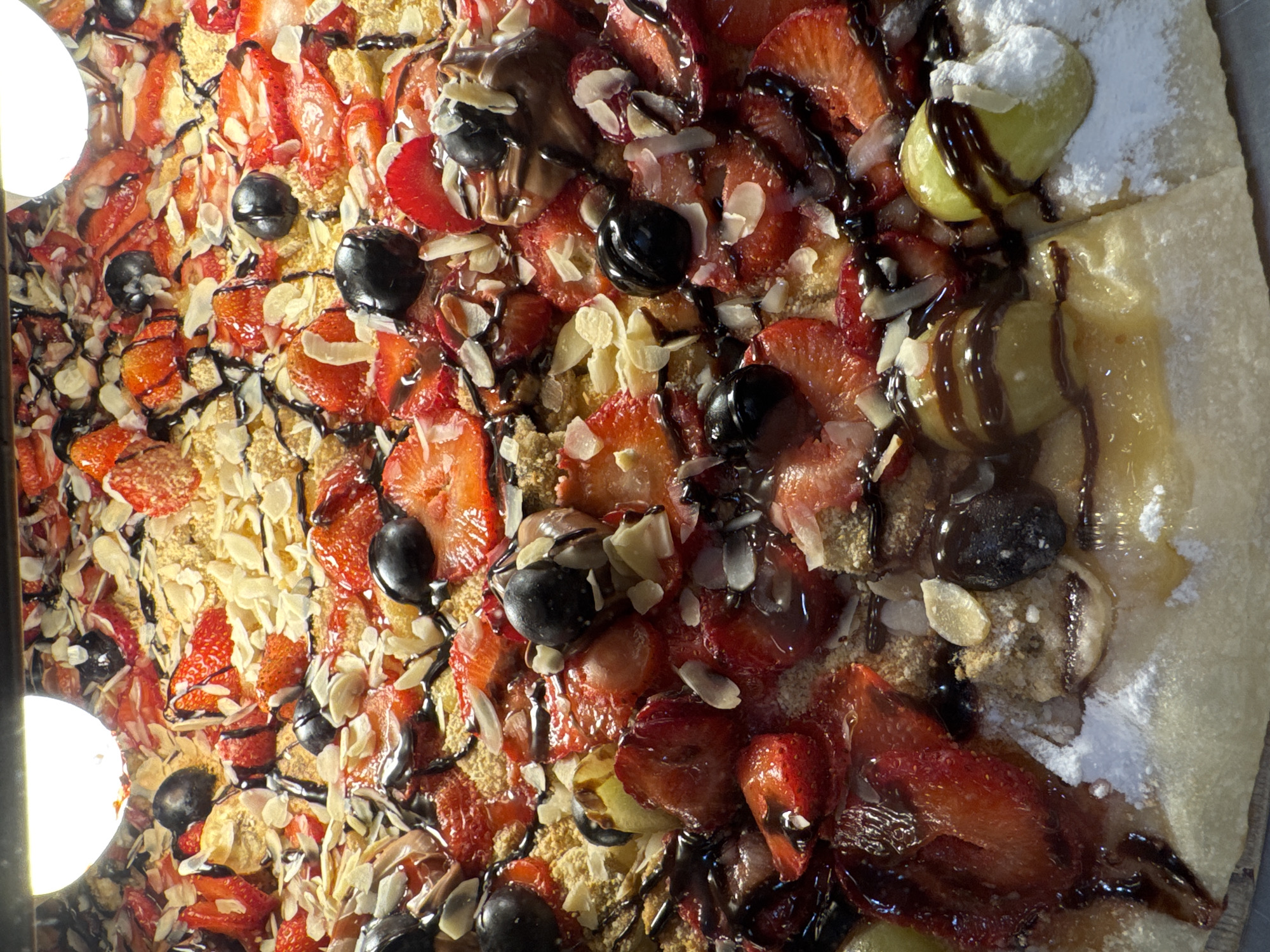 The image depicts a close-up of a dessert pizza topped with strawberries, blueberries, sliced almonds, chocolate sauce, and possibly grapes. The pizza appears to be cut into slices, with the crust visible along the edge. A portion of the pizza is dusted with powdered sugar. The lighting is slightly uneven, creating highlights on the toppings. The presence of this pizza was confirmed on location in the city of Budva, Montenegro.