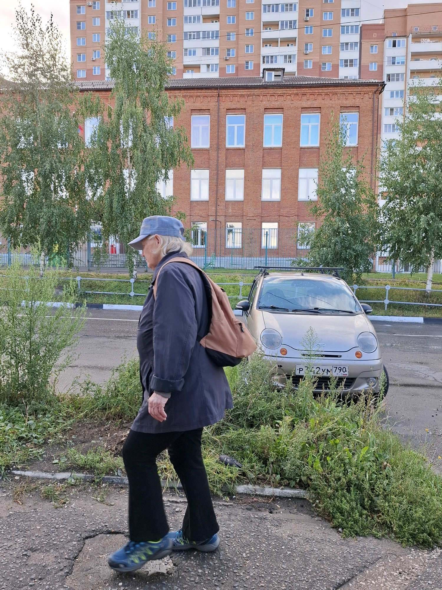 An individual wearing a blue cap, a dark blue coat, and black pants walks on a cracked asphalt path in front of a silver car. The individual is carrying a light brown backpack. Behind the individual and the car are buildings and trees. The car has a license plate that reads "P342YM 790 RUS". The ground is covered with gravel and patches of grass. The sky is overcast.