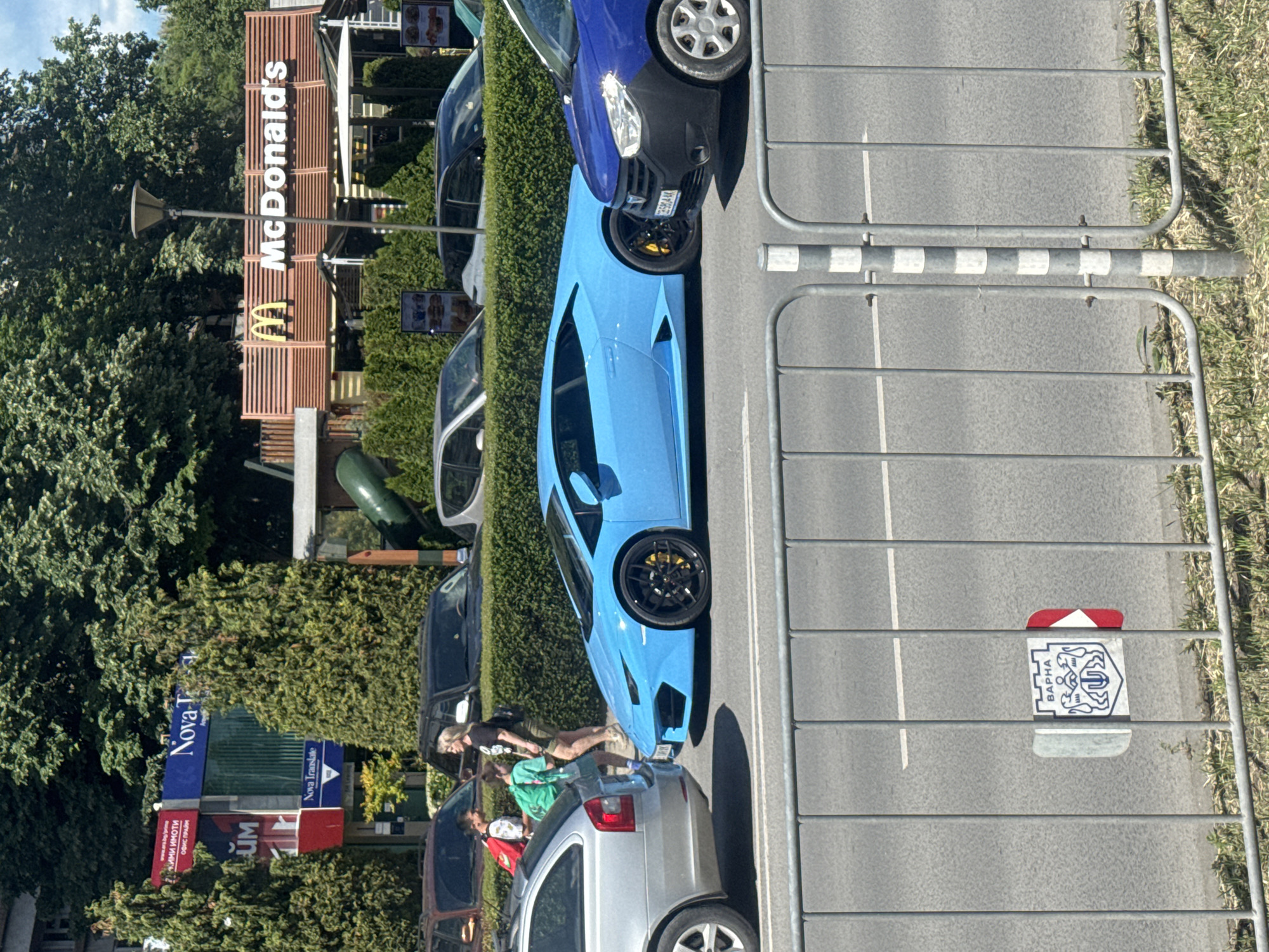 The image shows a street scene in Varna, Bulgaria. A bright blue Lamborghini is parked on the street. Next to it, a silver car is parked, near which two women and a child stand. Further down the street, there are several parked cars and a McDonald's restaurant. A Nova-T Translate sign can also be seen among the trees. The street is separated by a metal fence with a Varna city sign attached to it.