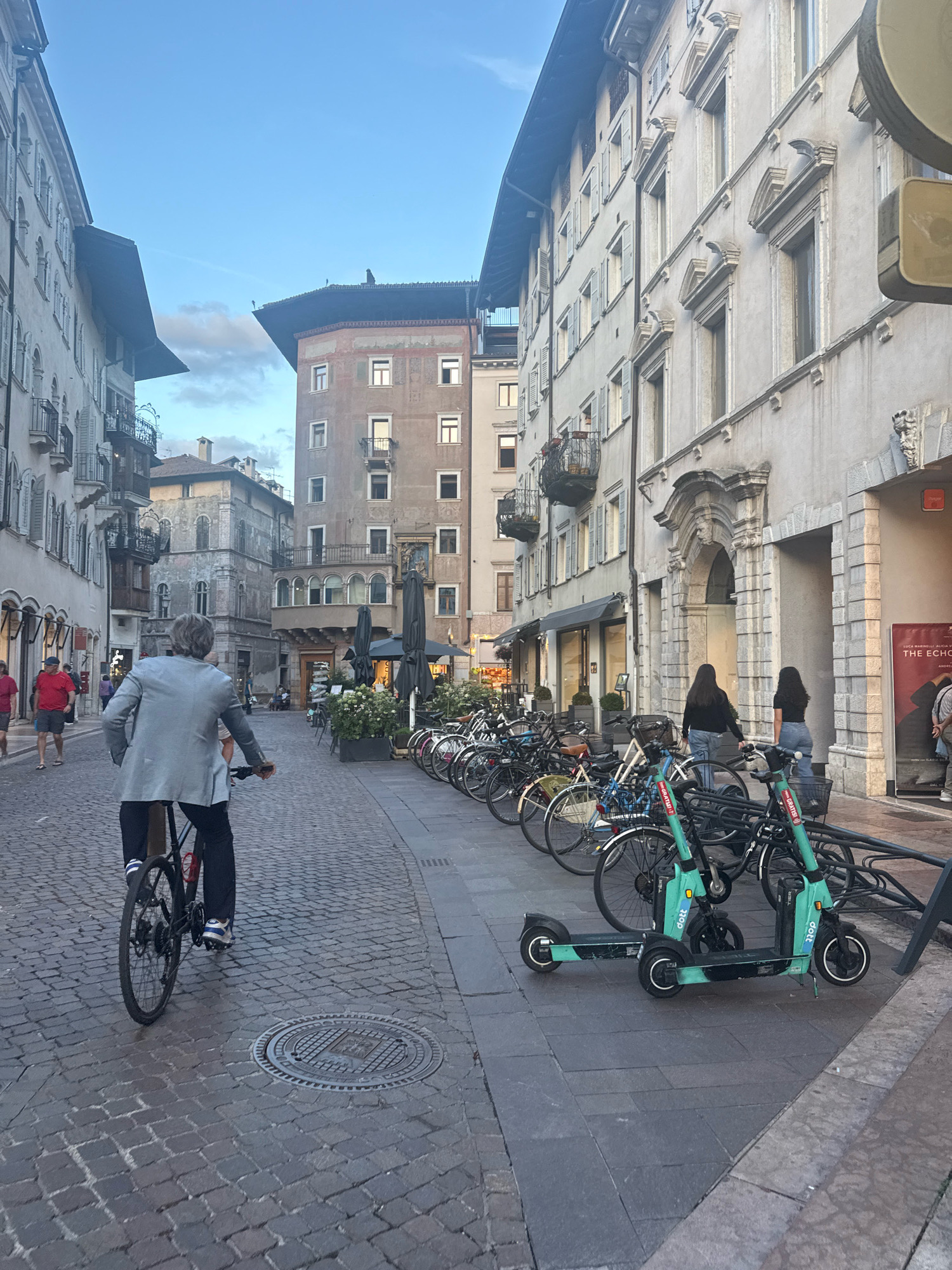 The image captures a street scene in Trento, Italy, characterized by its cobblestone pavement and historic architecture. In the foreground, a person is riding a bicycle, viewed from behind. The cyclist is wearing a light-colored blazer, dark pants, and striped socks with athletic shoes. To the right of the cyclist, several electric scooters are parked, some of which are bright green with "dott" branding. A row of parked bicycles is visible further down the street, adjacent to a building with an arched entryway and ornate architectural details.
The buildings lining the street exhibit traditional Italian architectural styles, with multiple stories, windows with shutters, and decorative elements. Several people are walking on the sidewalk in the background, and some are seated at outdoor tables, suggesting a commercial or residential area. Umbrellas are set up outside what appears to be a cafe or restaurant. The sky above is clear with a few wispy clouds, indicating it might be late afternoon or early evening. The overall impression is of a lively urban environment in a historic European city.