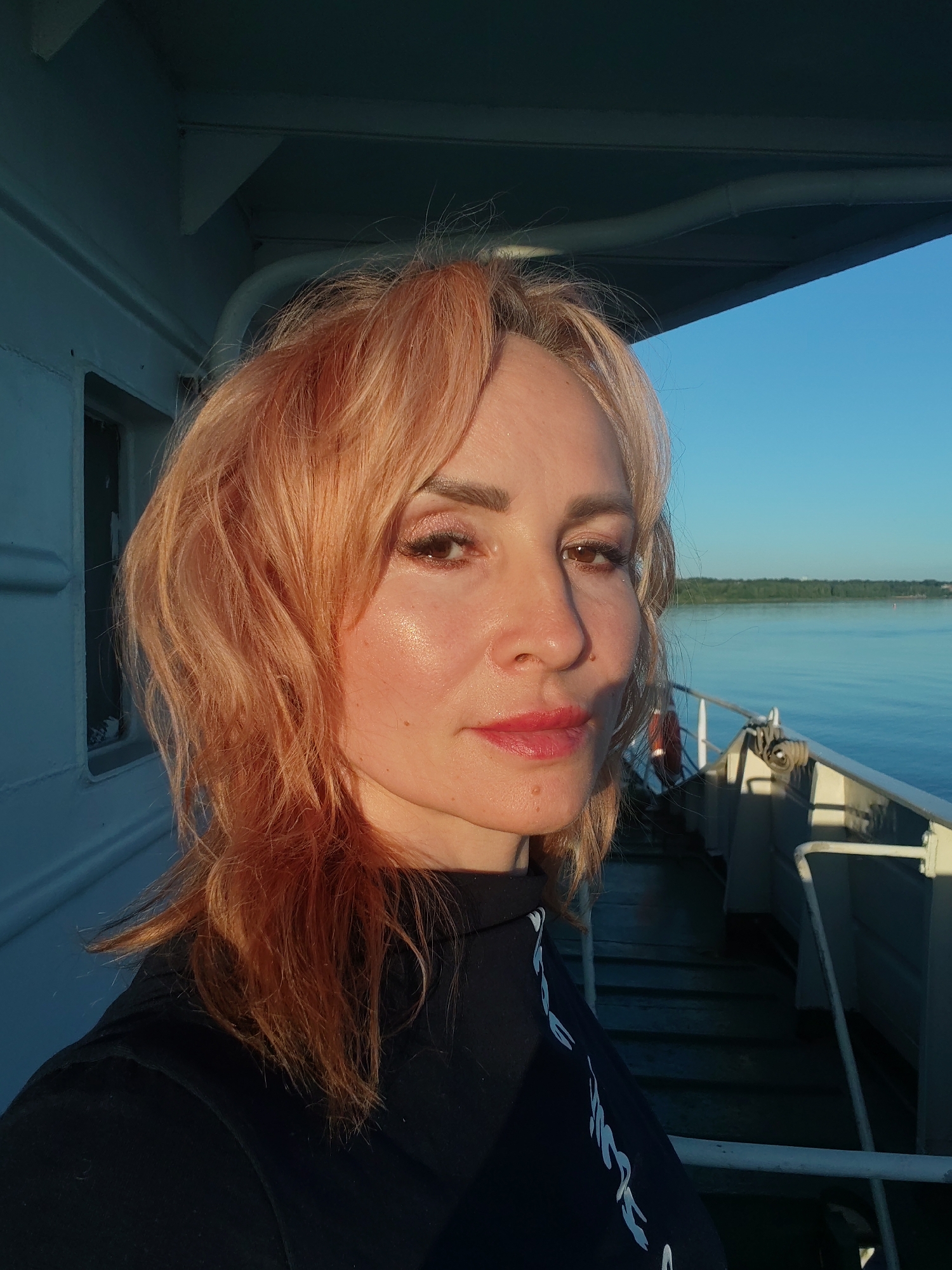 A middle-aged woman with reddish-blonde hair stands on the deck of a boat, possibly a ferry, with a clear blue sky and calm water in the background. She is wearing a black shirt. The boat appears to be docked or moving slowly, with railing and stairs visible on the deck. The lighting suggests it is either sunrise or sunset, casting a warm glow on her face and hair. The location is stated to be the city of Rybinsk, Russia.
