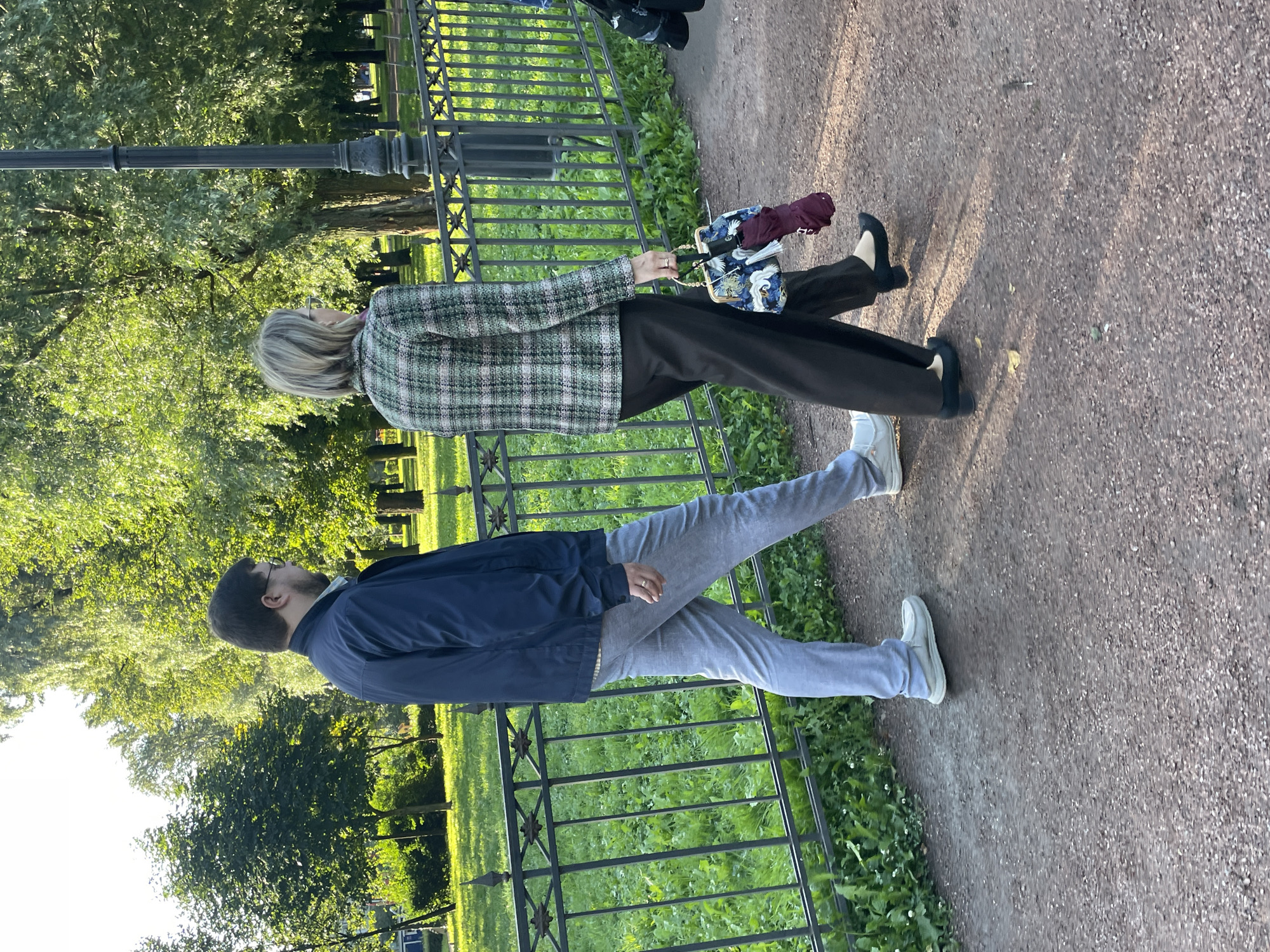 Two individuals are walking on a gravel path in a park. The person on the left is a male wearing a dark blue jacket, a light blue collared shirt, and light gray trousers. He has on light-colored slip-on shoes and glasses. The person on the right is a female wearing a checkered blazer, dark trousers, and black low-heeled shoes. She is carrying a handbag with a colorful pattern and a folded burgundy umbrella. They are walking side-by-side, facing away from the camera. Behind them, a black metal fence separates the path from a grassy area with trees. A lamppost is visible to the right. The scene appears to be in a city park, likely in Saint Petersburg, Russia, given the architectural style of the lamppost and the presence of mature trees and manicured greenery often found in the city's public spaces. The lighting suggests it is daytime, possibly late afternoon, with the sun casting shadows on the path.