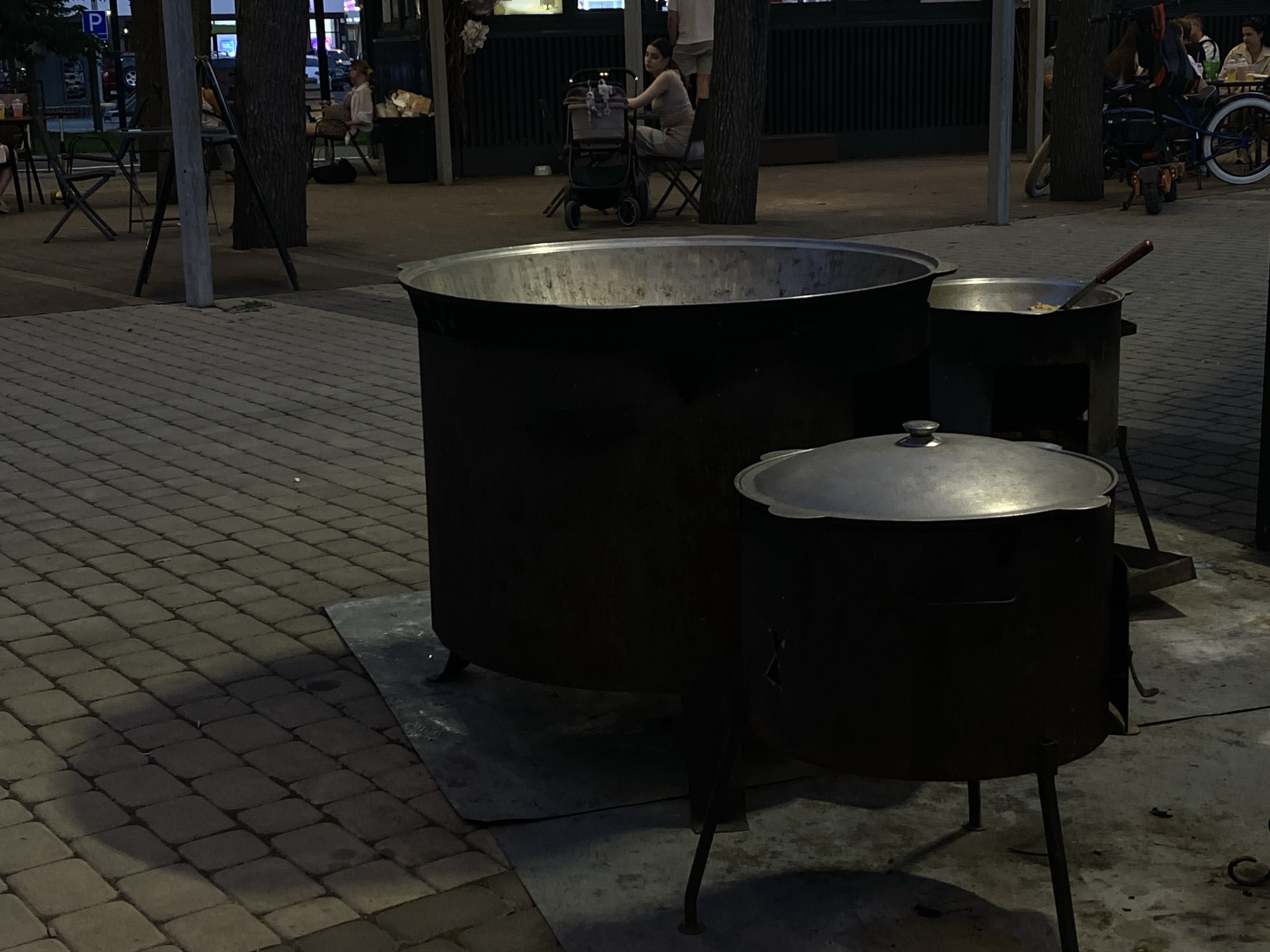 In an outdoor, paved area that appears to be a public space or park in Krasnodar, Russia, two large, dark metal cooking vessels are positioned on metal stands. The larger of the two is to the left and is open at the top, revealing its interior. To its right, a smaller, lidded vessel sits on a stand with an opening for a fire below. A cooking utensil, possibly a ladle or large spoon with a reddish-brown handle, is visible resting in the opening of a metal structure beside the smaller pot, suggesting active food preparation. In the background, trees line the area, and several people are visible sitting at tables and a baby stroller is present. A bicycle with white-rimmed wheels is also parked nearby. The lighting suggests it is either dusk or a shaded area.