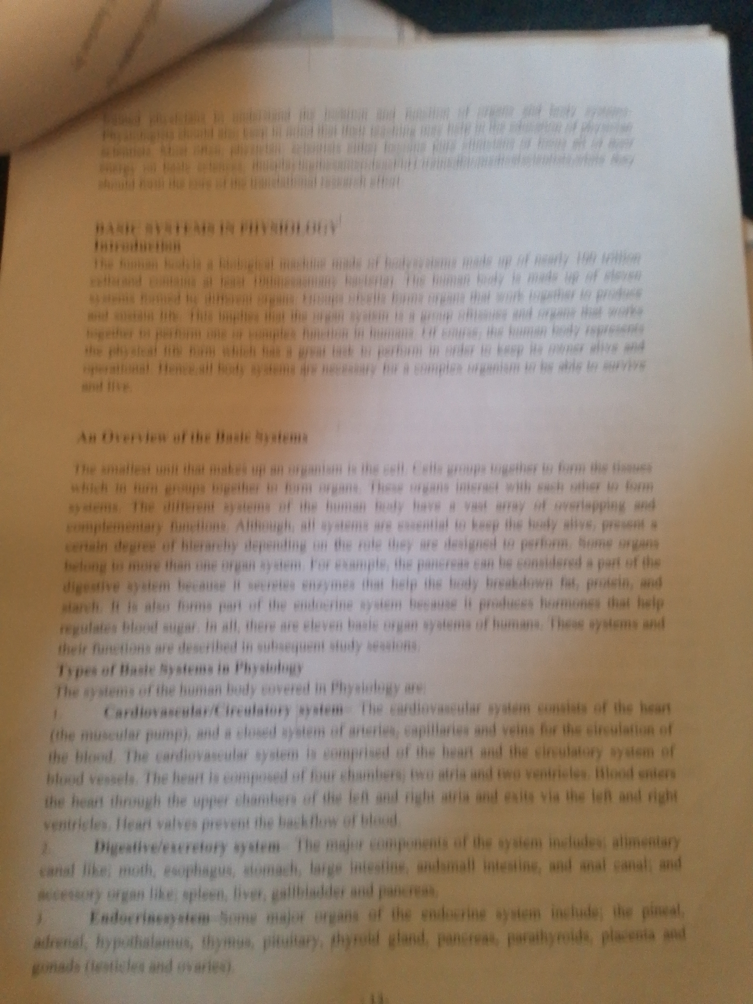This media file is a photograph of an open book, specifically a page from a textbook discussing the basic systems of human physiology. The text is printed in black on white paper. The book appears to be lying flat, with the left side of the page slightly curled upwards. The lighting suggests an indoor setting, possibly a room with ambient light. There are no people visible in the image.
The main subject is the text itself, which details the introduction to basic systems in physiology, an overview of these systems, and then begins to list and describe specific systems. Visible text includes headings such as "BASIC SYSTEMS IN PHYSIOLOGY," "Introduction," "An Overview of the Basic Systems," and "Types of Basic Systems in Physiology." It then lists and describes the "Cardiovascular/Circulatory system," "Digestive/excretory system," and "Endocrinesystem."
The image is slightly blurry and taken from an angle, suggesting it was captured with a handheld device. The focus is on the printed content, with no other objects or background elements clearly discernible. The page number "13" is visible at the bottom center of the page. The context provided places the location as Jalingo, Nigeria, but there are no visual cues within the image itself to support or contradict this. The content of the book relates to biological science and human anatomy.