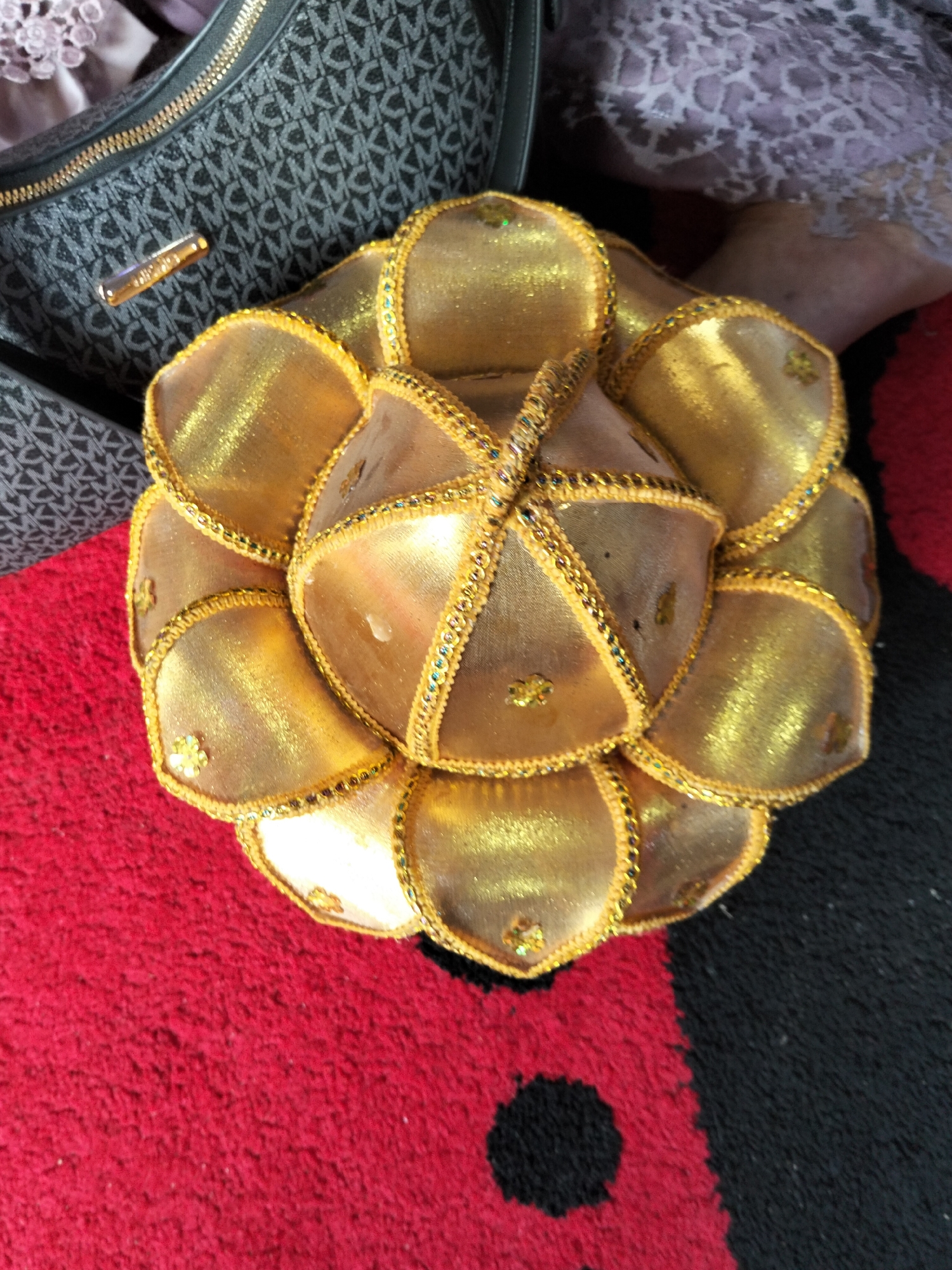 The image shows a close-up, overhead view of a gold, petal-shaped decorative item. The item is placed on a red carpet with a black and red striped pattern. To the upper left of the decorative item is a black handbag with a gray MK logo pattern. A small, gold-colored plaque with the inscription "MK" is visible on the handbag. In the upper right corner, a portion of a person's leg is visible, wearing a light purple fabric with an intricate pattern. The scene is indoors, and the lighting suggests natural light coming from above. The image does not contain information about the city of Cottrueng, Indonesia.