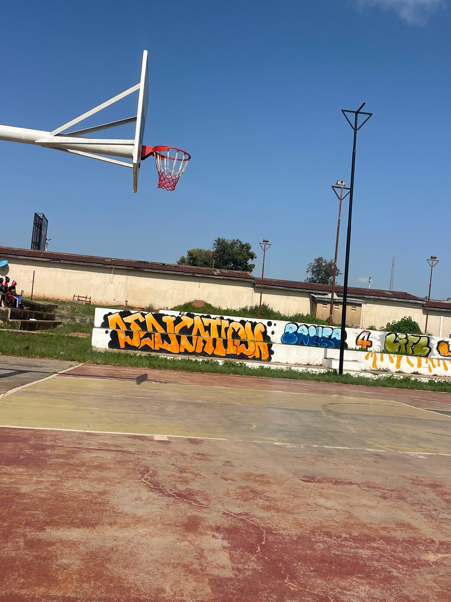 An outdoor basketball court with a weathered red and yellow surface is depicted under a clear blue sky. A white basketball hoop with a red net is visible on the left side of the frame. A long, beige building with a corrugated roof runs along the background. A low concrete wall separating the court from the building is covered in graffiti. The graffiti features large, colorful lettering, predominantly in orange, blue, and green. To the left, a set of concrete bleachers is partially visible, with several young individuals seated on them. Two tall, dark lampposts stand near the wall, one in the foreground and another further back. The scene appears to be in a somewhat urban or semi-urban environment in Jos, Nigeria, characterized by the presence of the basketball court, the building, and the graffiti.
