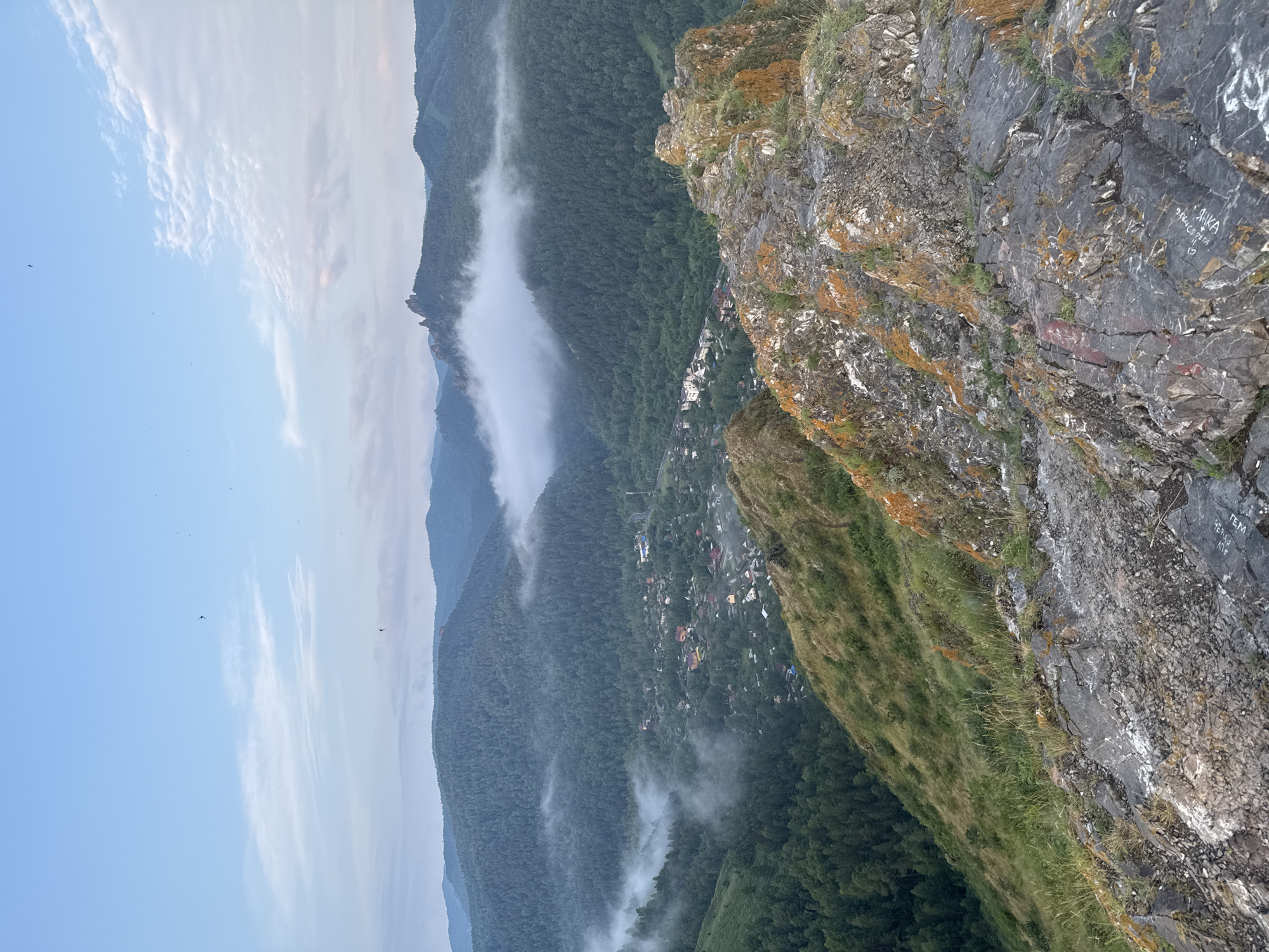 The image captures a panoramic view from a rocky outcrop overlooking a valley with a village nestled among forested hills. The foreground is dominated by the textured surface of the rocky cliff, with patches of moss and lichen in shades of orange and green. Beyond the immediate foreground, the landscape descends into a lush green valley filled with dense conifer forests. A small village is visible in the valley, with scattered houses and buildings. Wisps of white fog or mist are present, clinging to the slopes of the hills and partially obscuring the distant peaks. The sky above is a pale blue with some scattered clouds, suggesting either early morning or late evening. Several small birds are visible in flight against the sky. The overall impression is one of natural beauty and tranquility. This scene is characteristic of the area around Krasnoyarsk, Russia, specifically views from the Stolby Nature Reserve, known for its dramatic rock formations and extensive taiga forests overlooking the Yenisei River and surrounding settlements.