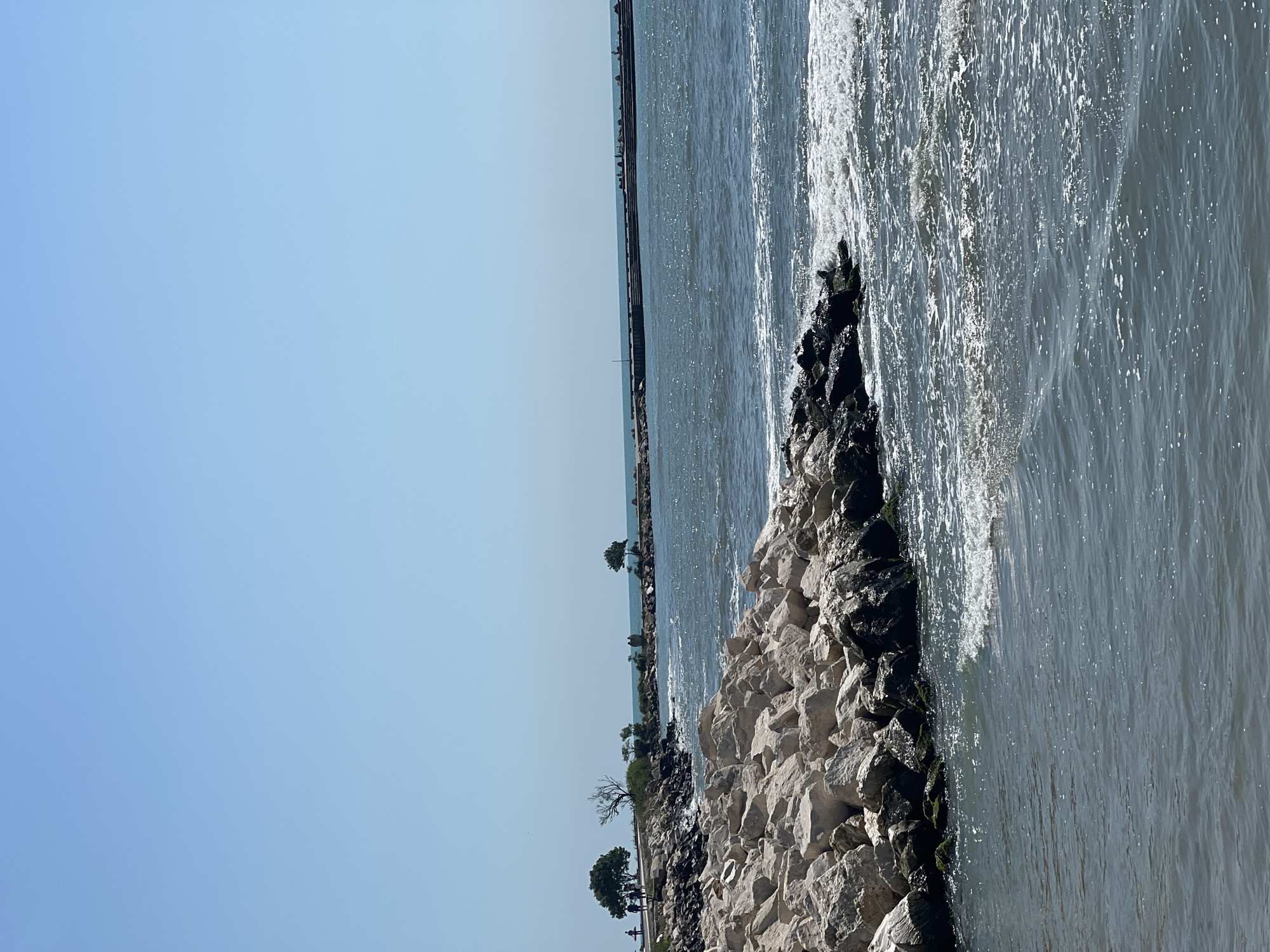 The image presents a bright, sunlit coastal scene, likely a breakwater or rocky shoreline, under a clear blue sky, characteristic of Varna, Bulgaria.
The upper portion of the frame is dominated by a vast, cloudless, light blue sky, indicating fair weather. Below the horizon, which cuts horizontally across the middle of the image, lies the sea and a rocky barrier.
The lower-left side of the image features a substantial rocky revetment or breakwater. It is composed of numerous stones of various sizes, ranging from light grey to darker tones, with some exhibiting traces of green moss or algae. This rocky formation extends diagonally from the lower-left foreground towards the mid-ground, separating the land from the water.
To the right of this rocky barrier, the sea stretches out. The water's surface is choppy with ripples and reflects bright sunlight, creating shimmering patterns. Visible waves are breaking against the rocks, generating white foam.
In the far distance, along the horizon line, a long, narrow structure, likely a pier or a distant section of a breakwater, extends into the sea. Small, indistinct figures, potentially people, are barely discernible on this distant structure and along a narrow strip of land behind the main rocky barrier, where a few small, sparse trees are also visible.
The scene suggests a pleasant, calm day, likely during daytime, by the Black Sea. There is no visible text in the image. No specific human activity is clearly prominent, though the distant figures imply leisure or observation.