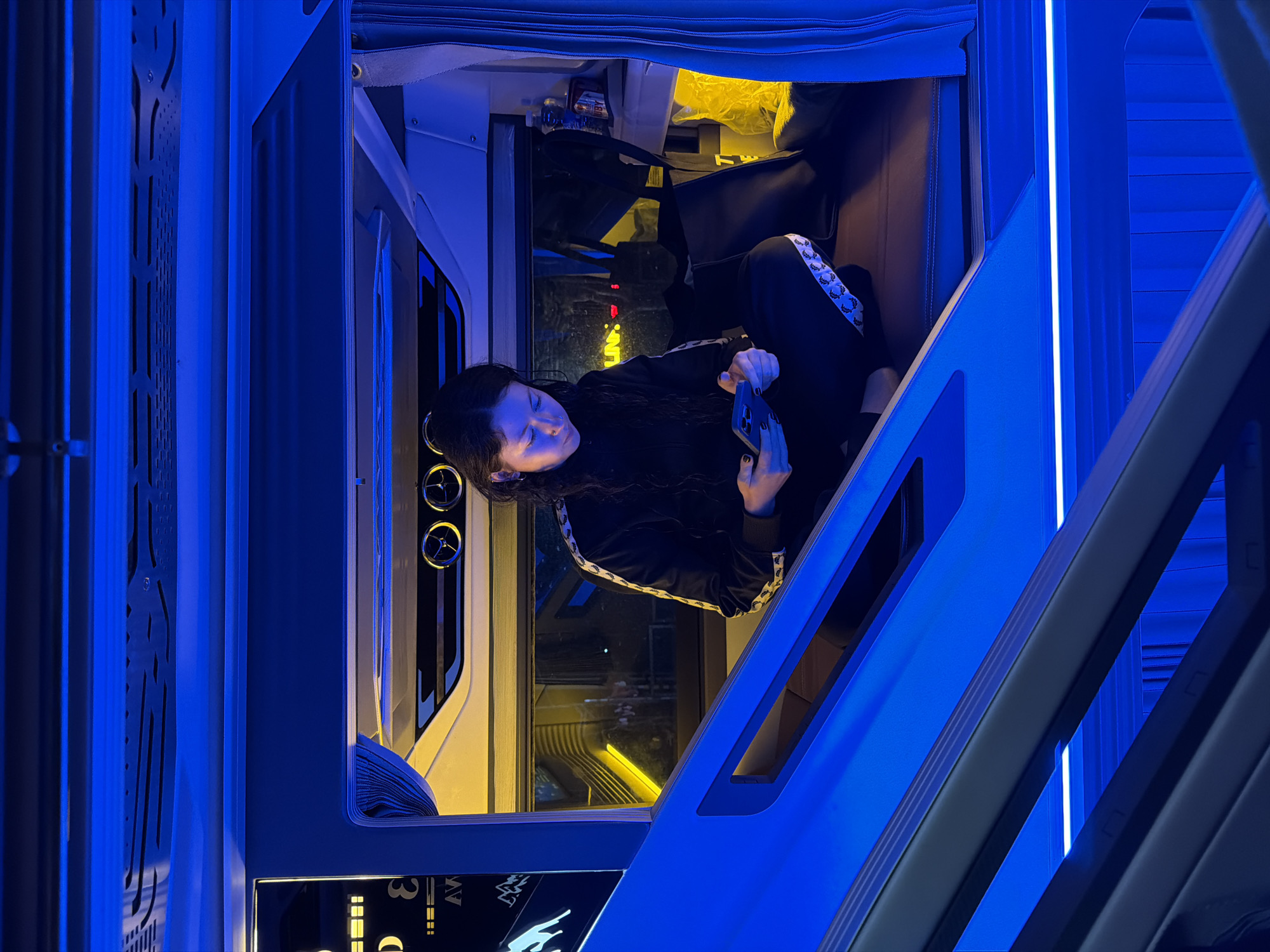 The image captures a young woman inside a modern, futuristic-looking capsule or compartment, likely a sleeping pod in a hostel or a private bunk in a sleeper train, in Đà Nẵng, Vietnam. She has dark hair and is wearing a black track jacket with white and black striped trim on the sleeves, and dark pants. She is positioned inside the compartment, reclining or lying down, and is focused on a dark-colored smartphone held in her hands.
The interior of her compartment is softly illuminated by a warm yellow light, creating a cozy contrast with the striking blue LED lighting that dominates the surrounding structure. Above her head, two circular vents or light fixtures are visible. The exterior structure features sleek, geometric lines and perforated patterns, all bathed in an intense blue glow. The overall impression is one of a compact, well-lit private space within a larger, modern vehicle or building.
Visible text on a panel below her compartment includes parts of "3" and "AV," along with a symbol that appears to be a leaping animal. The patterned trim on her clothing also suggests a repeating brand logo. The scene takes place at what appears to be nighttime or in an interior setting reliant on artificial light. The atmosphere is calm, with the woman engaged with her phone in a private, enclosed setting.