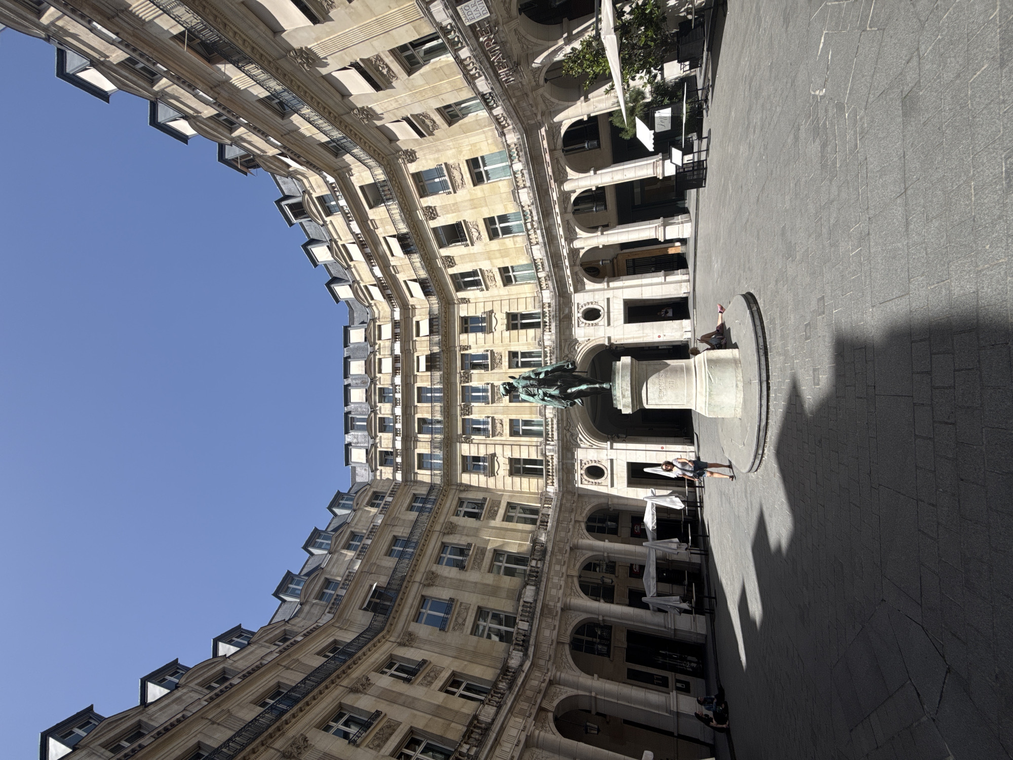 The image captures a courtyard in Paris, France, enclosed by a grand, cream-colored building with numerous windows and wrought-iron balconies. The sky is a clear blue, visible through the building's open structure. In the center of the courtyard stands a statue atop a pedestal, casting a long shadow across the gray, paved ground. Several people are in the scene, including a woman and a child near the statue, and another person walking in the lower right. Tables and chairs are arranged along the building's arches, suggesting a cafe or seating area. The building architecture features arched entryways and detailed stonework.
