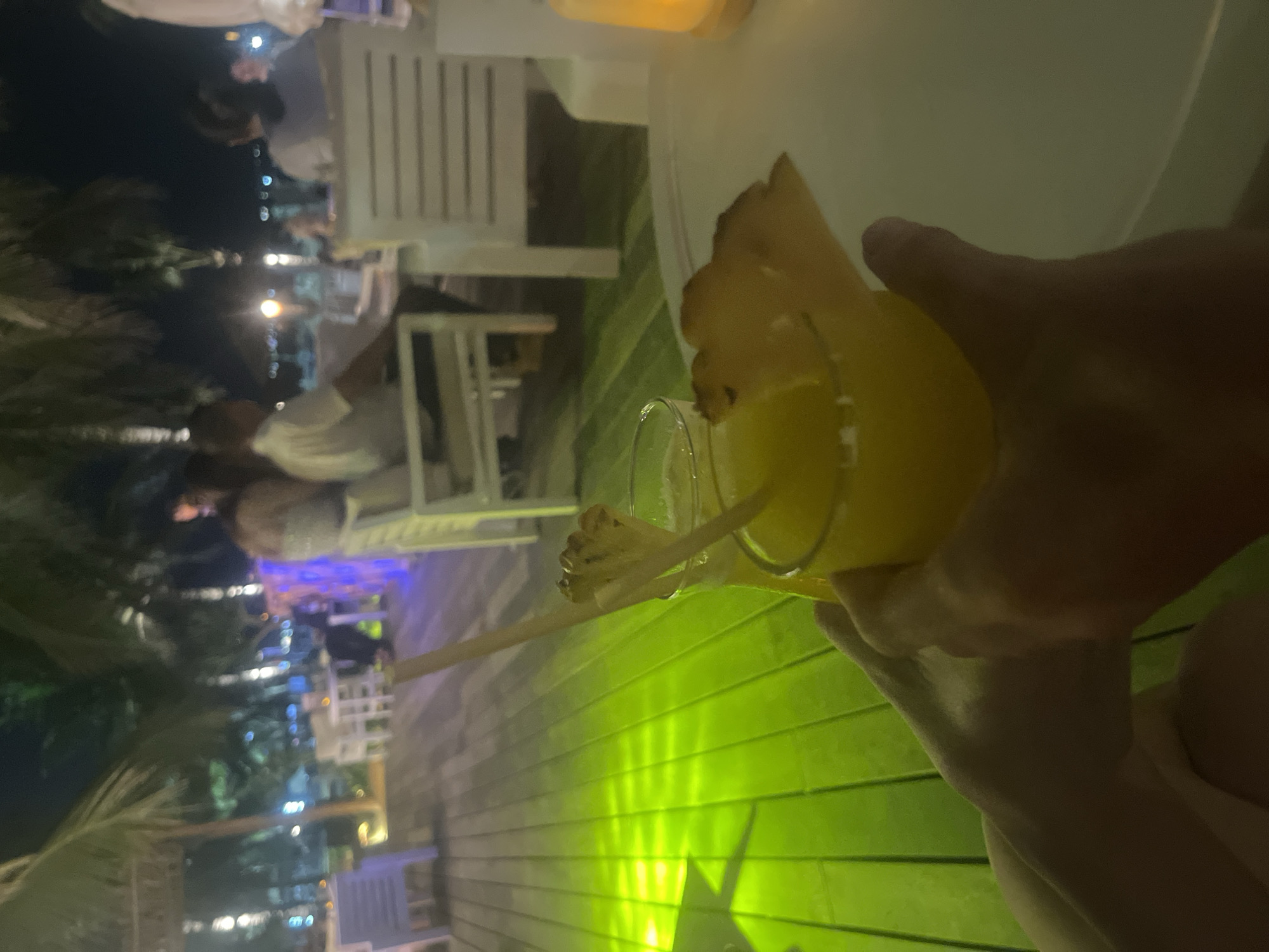 At night, at a beachside restaurant in Da Nang, Vietnam, two people are holding drinks. The person in the foreground is holding a glass of yellow liquid with a pineapple garnish and a straw. The other person's hand is also visible, holding a similar drink. The restaurant has a wooden deck and white chairs. Palm trees and other vegetation are visible in the background, along with the lights of other patrons. The overall atmosphere is relaxed and festive.