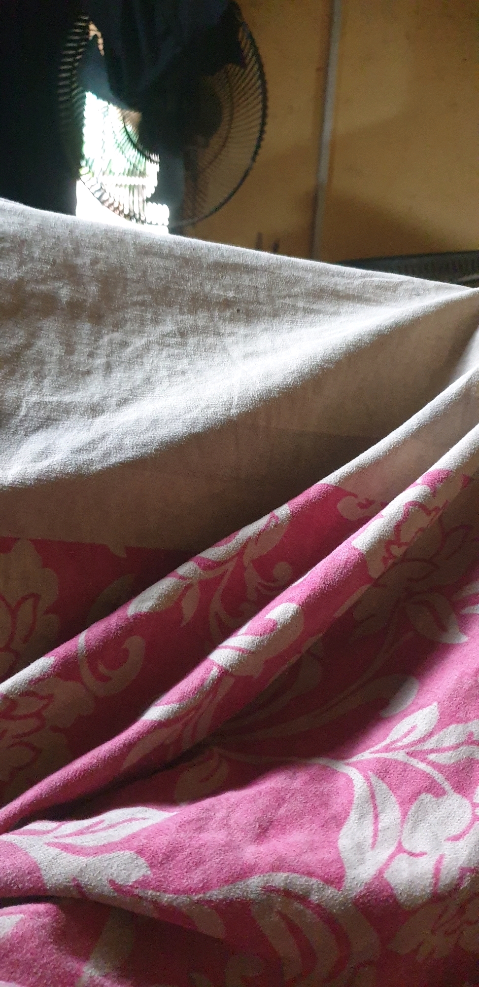 The image shows a close-up of a bed with crumpled sheets in a room. The sheets are a light beige and a pink with a floral pattern. In the background, partially visible, is a dark electric fan with its blades obscured by the sheets and lighting. The wall behind the fan is a plain, warm yellow color. The lighting suggests an indoor setting, possibly during the day with light coming from a window or opening behind the fan. The context of Sabon Tasha, Nigeria, indicates this is likely a domestic scene in a residential area.