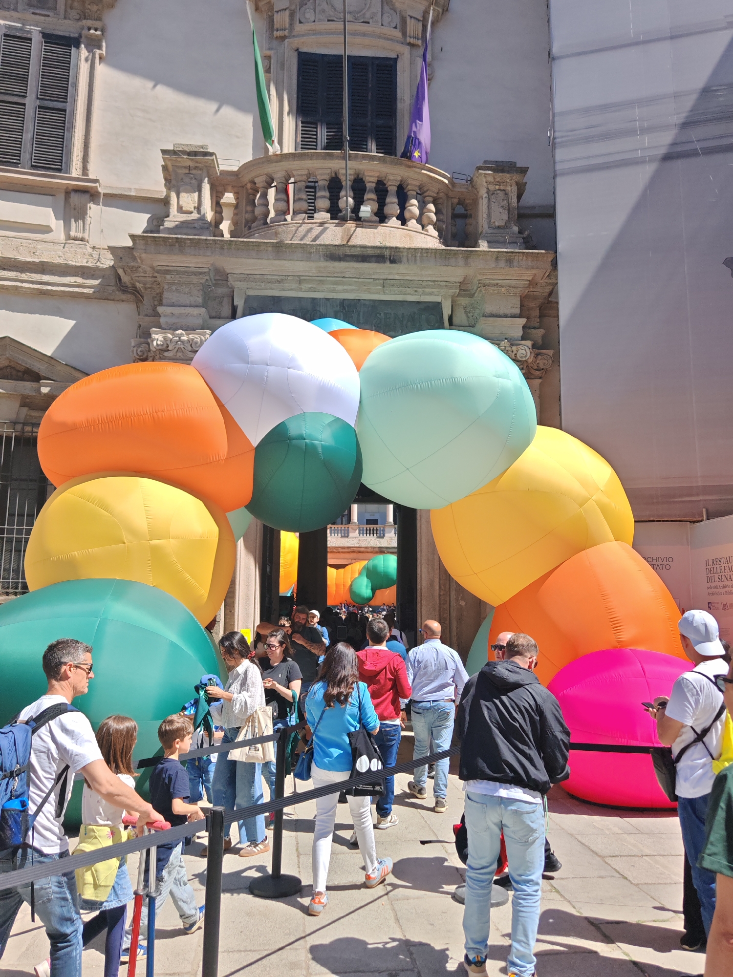 Here is a detailed description of the image:
**General Scene:**
The image shows a crowd of people lined up to enter a building, likely an historical site or venue. The entrance is adorned with a large, colorful arch made of inflated, spherical balloons. There are multiple people of different age groups scattered throughout the scene.
**Specific Subjects and Objects:**
1. **Balloons:** A prominent feature is the arch made of large inflated balloons in multiple colors including white, orange, yellow, green, pink, and teal.
2. **Building:** The building appears to be of classical architecture. There are shutters on the windows, a balcony above the entrance, and two flags are seen on the building's facade.
3. **Crowd:** The group is composed of various ages from children to adults, standing in a line that is managed by stanchions and rope barriers.
4. **People:** Several individuals are visible. The crowd contains men, women, and children. A child is on a scooter, and various individuals are holding bags or using phones.
5. **Signage:** There is a sign indicating something about restoration work being done. It says something about "Archivio Tato" and "Il Restauro delle Fac...del Senato."
**Actions and Interactions:**
* People are queued up, presumably to enter the building.
* Some people are looking at their phones.
* A father and child are walking together, a family group is visible.
* Some people are talking amongst themselves.
**Environment:**
* The scene is well-lit, suggesting it is daytime and the weather is likely sunny.
* The ground is paved, with visible lines/joints in the paving.
* The scene appears to be in a urban area, given the type of building and surrounding infrastructure.
**Location:**
Based on the signage visible in the image, it is likely in or near the Archivio Storico del Senato (Historical Archive of the Senate), which is the building in the background.