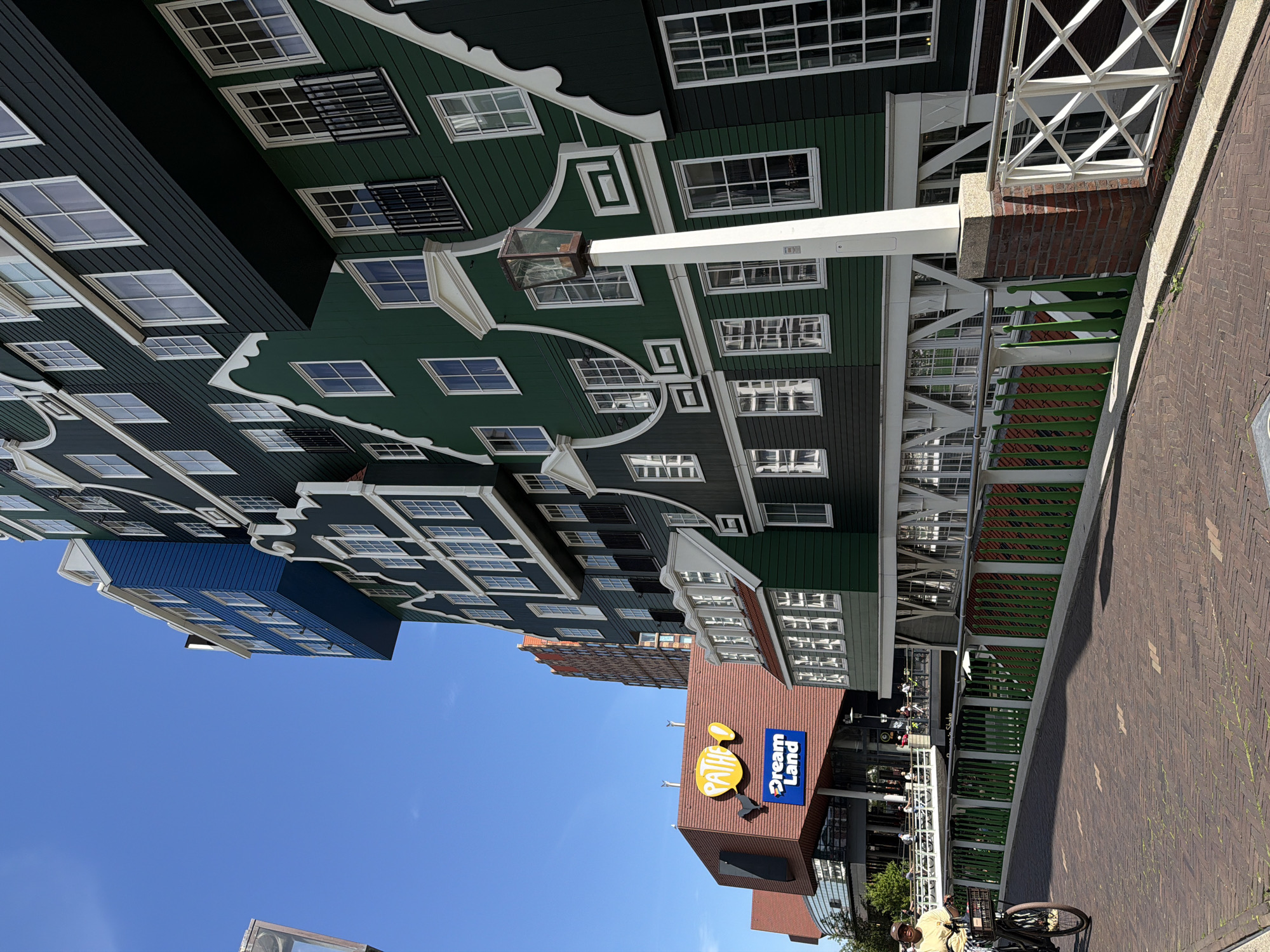 In the city of Zaandam, Netherlands, a cyclist rides on a brick-paved street in front of a distinctive green building complex with a blue section. The building exhibits a series of stepped gable facades, a traditional Dutch architectural style, with numerous windows. To the left of the green building is a red structure with a sign that reads "Pathe" and "Dream Land" in blue letters. The sky is clear and blue. The cyclist is wearing a yellow shirt and a hat, and is on a bicycle with a basket. The scene captures a sunny day with shadows cast on the street.
