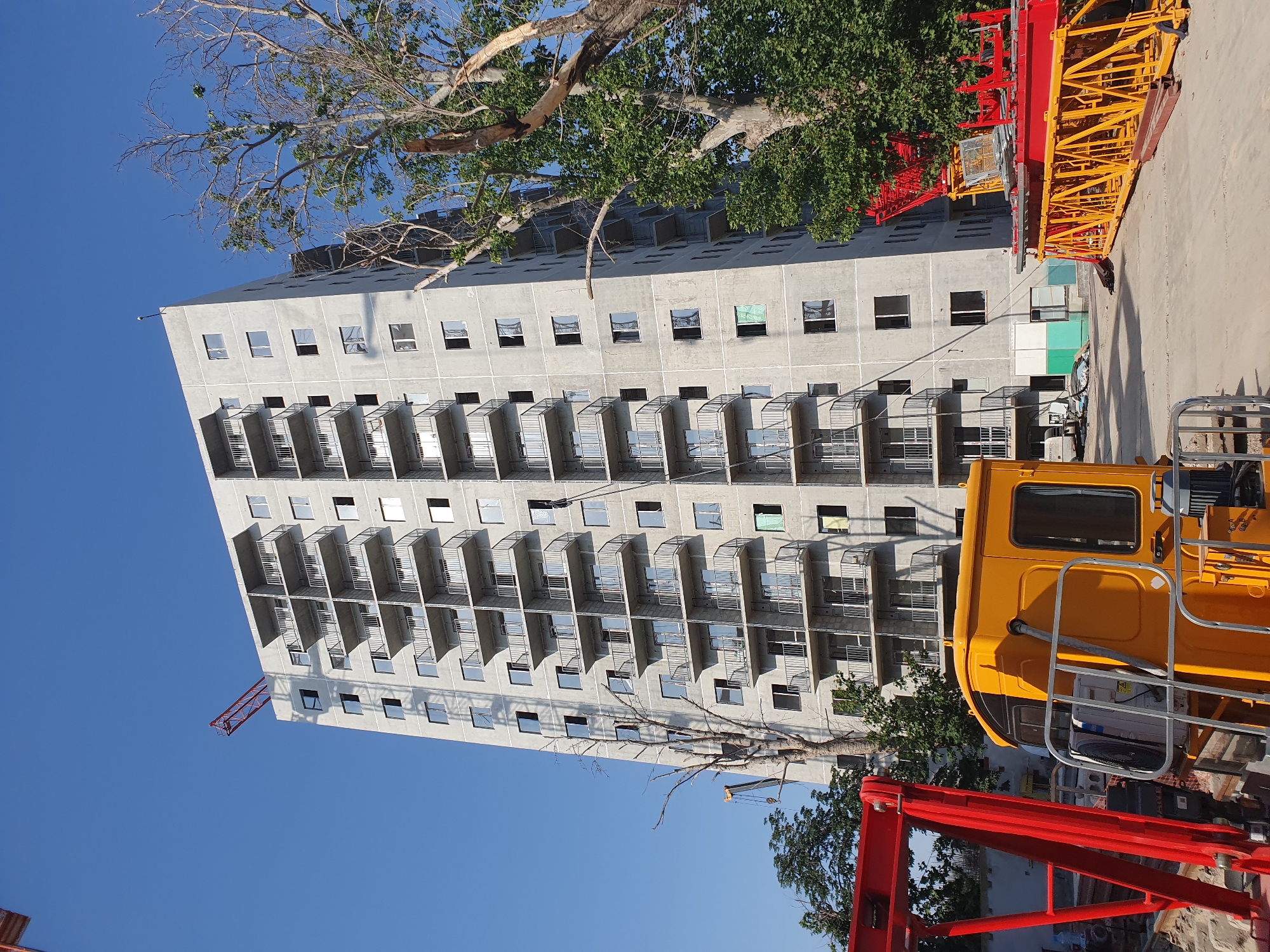The image depicts a large, unfinished apartment building in Tashkent, Uzbekistan. The building is constructed of concrete and has multiple balconies with metal railings. Windows are visible on each floor, and some appear to be open. Construction equipment, including a red crane boom and a yellow construction vehicle, is present on the ground. Several trees, some appearing dead or damaged, are also visible around the building. The sky is clear and blue, suggesting a sunny day. The overall scene suggests an active construction site in an urban environment.