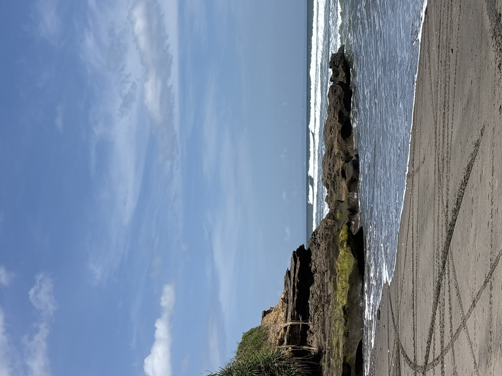 The image depicts a coastal scene in Kerambitan, Indonesia. The sky is blue with wispy clouds. The ocean is visible with small waves breaking near the shore. Rocky cliffs with some vegetation are located along the edge of the beach. The sand is dark in color with several tire tracks visible. There are no people depicted in the scene.