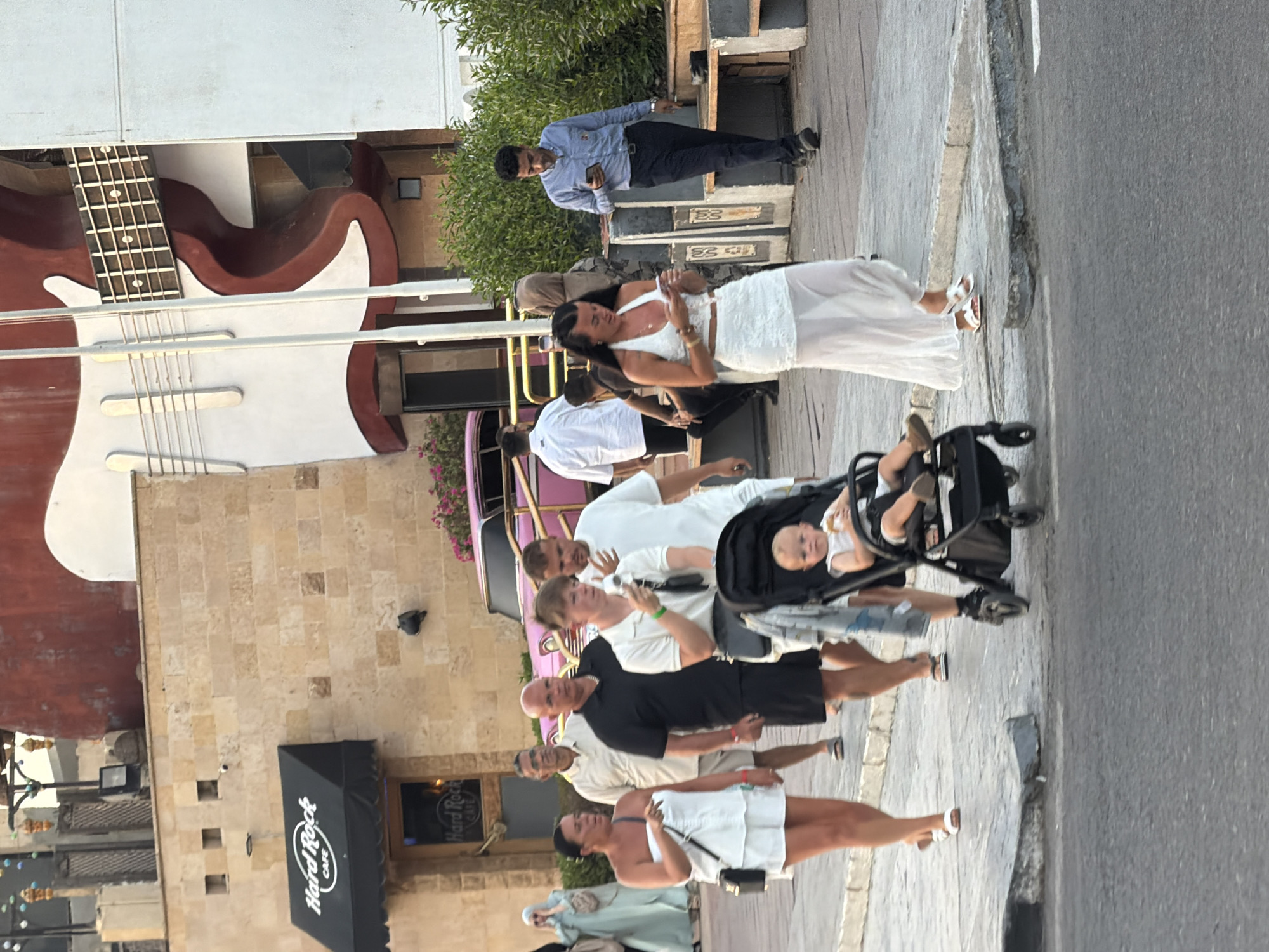 The image depicts a street scene in Sharm El Sheikh, Egypt, in the vicinity of a Hard Rock Cafe. Several individuals are walking on a paved sidewalk. In the foreground, a woman in a white strapless top and shorts walks with a stroller containing a baby. To her right, a woman in a white halter top and a long white skirt walks while looking at her phone. Behind them, a man in a blue shirt and dark pants also looks at his phone. Further back, a group of people are walking together, including a man in a black t-shirt and shorts, a man in a light-colored polo shirt, and a man in a white t-shirt. In the background, the iconic Hard Rock Cafe building is visible with its distinctive awning. To the right of the Hard Rock Cafe, a large, stylized guitar sculpture adorns the building. A pink vintage car is also partially visible in the background. The environment suggests a warm, possibly evening, setting with buildings and vegetation lining the street.