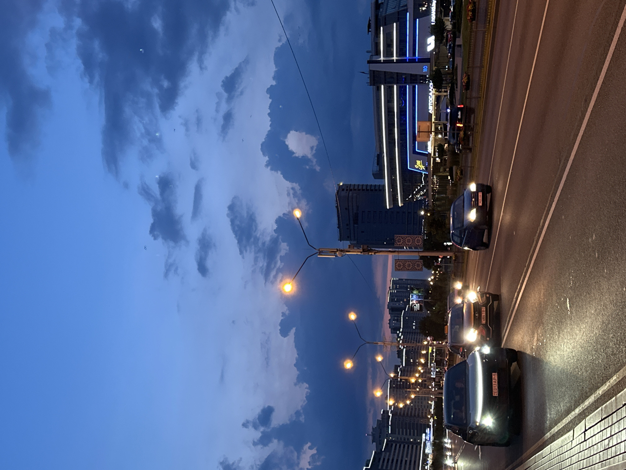A vertical, medium shot captures a city street at dusk, featuring a twilight sky with dark, textured clouds. Several cars are visible on the multi-lane road, their headlights and taillights illuminating the asphalt. The foreground shows the edge of the road with a white tiled border. In the mid-ground, streetlights with multiple bulbs cast a warm glow. Further back, a line of modern buildings stretches across the frame, some with illuminated windows and neon signage, suggesting a commercial or entertainment district. The overall atmosphere is that of a city transitioning from day to night, with artificial lights beginning to dominate the scene.