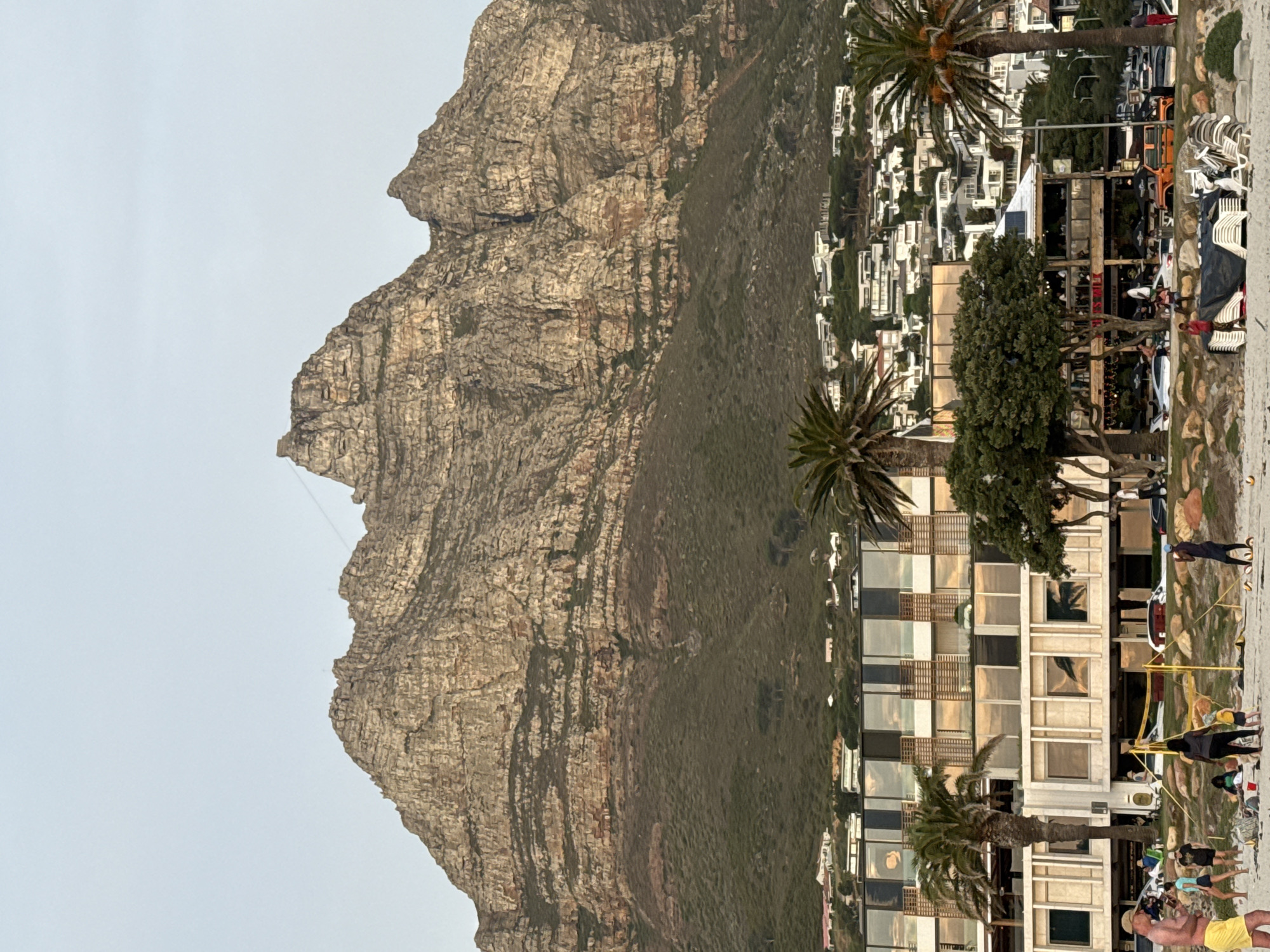 This image shows a view of Cape Town, South Africa. Prominently featured is a large, rugged mountain with a rocky face and sparse vegetation, likely Lion's Head or part of the Twelve Apostles range. Adjacent to the mountain, closer to the viewer, are buildings and palm trees suggesting a coastal urban environment. The lower portion shows signs of beach activity; people are visible interacting and walking on a beach-like area in front of buildings. There are also scattered objects such as chairs and structures that appear to be commercial establishments along the waterfront. The sky is overcast.