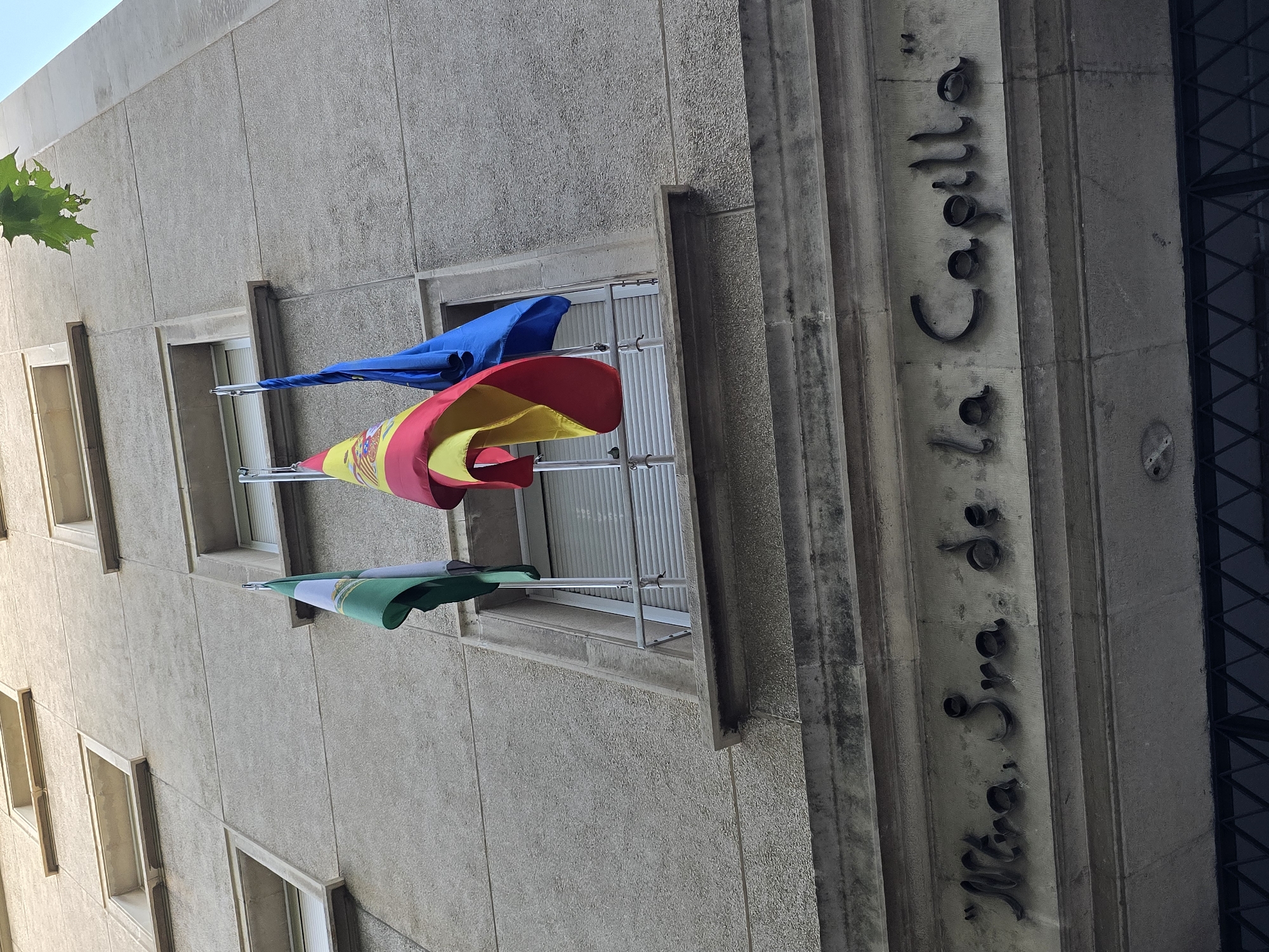 A low-angle, full shot captures the exterior of a building with a textured, light gray concrete facade. Three flags are mounted on poles outside a window on the second story. From left to right, the flags are green, white, and green with a crest; the Spanish flag, with red and yellow stripes and a crest; and a solid blue flag, likely the European Union flag. The flags are partially obscured by the building's architecture. Below the flags, a stone ledge juts out. Further down, an inscription in dark, raised metal lettering reads "Ntra. Sra. de la Capilla". The inscription is centered and spans across the facade. Below the inscription, a dark, metal-grated window or entrance is visible. A green leaf from a tree hangs in the upper right corner of the frame, silhouetted against a bright sky. The image has a slightly desaturated, muted color palette.