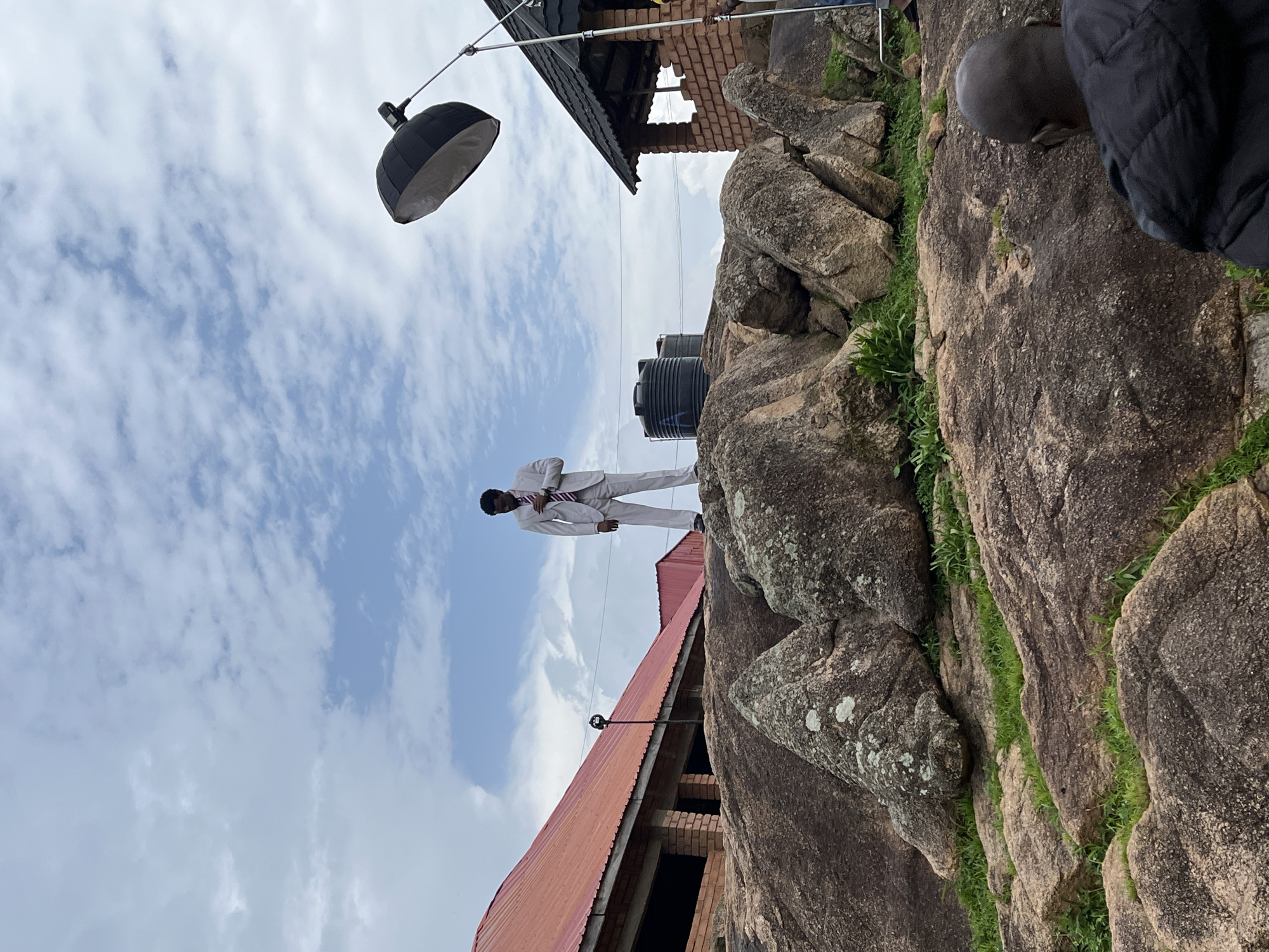 A person in a light-colored suit stands on a rocky outcrop against a cloudy sky. The person is leaning forward, with one leg extended. To the left of the person is a red-roofed building under construction, with exposed brickwork. Above and behind the building, a large black water tank is visible. A studio light on a stand is positioned near the building. The scene appears to be outdoors, with a natural rocky environment. The sky is partly cloudy, suggesting daytime. The rocky terrain and the building under construction might indicate a somewhat remote or developing area. The overall mood is somewhat formal due to the subject's attire, contrasted with the rugged natural setting. There is no visible text in the image.