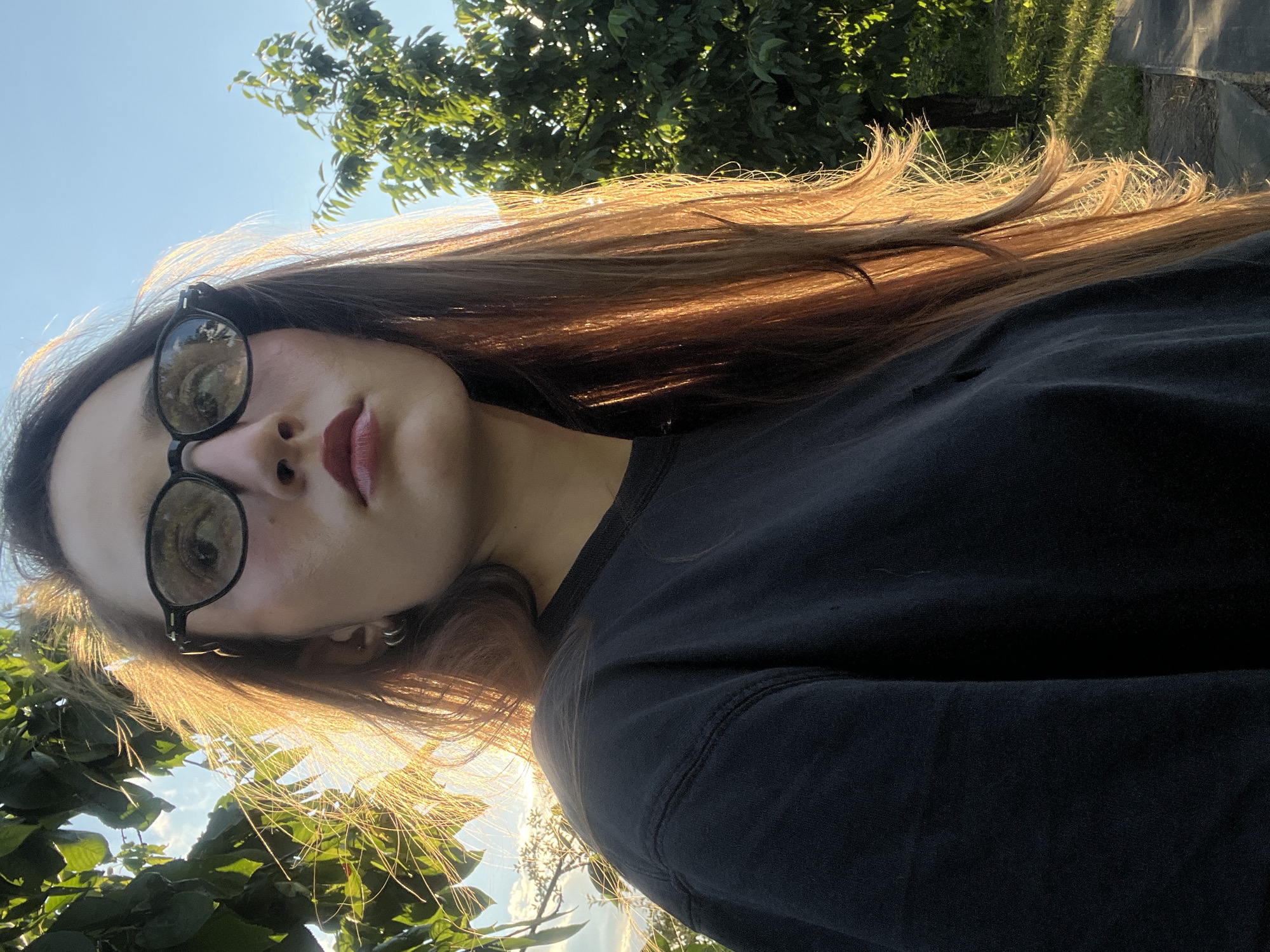 A young woman is taking a selfie outdoors, with the camera angled upwards. She is wearing round, dark-rimmed glasses and a dark t-shirt. Her long, light brown hair is catching the sunlight, appearing to glow. Her lips are painted a deep red.
The background consists of green foliage from trees and a bright blue sky. The lighting suggests it is either early morning or late afternoon, with the sun low in the sky and casting a warm glow. The overall mood appears relaxed and natural, with the woman's gaze directed towards the camera. There is no visible text in the image. The setting seems to be a garden or a park, with lush greenery and ample natural light.